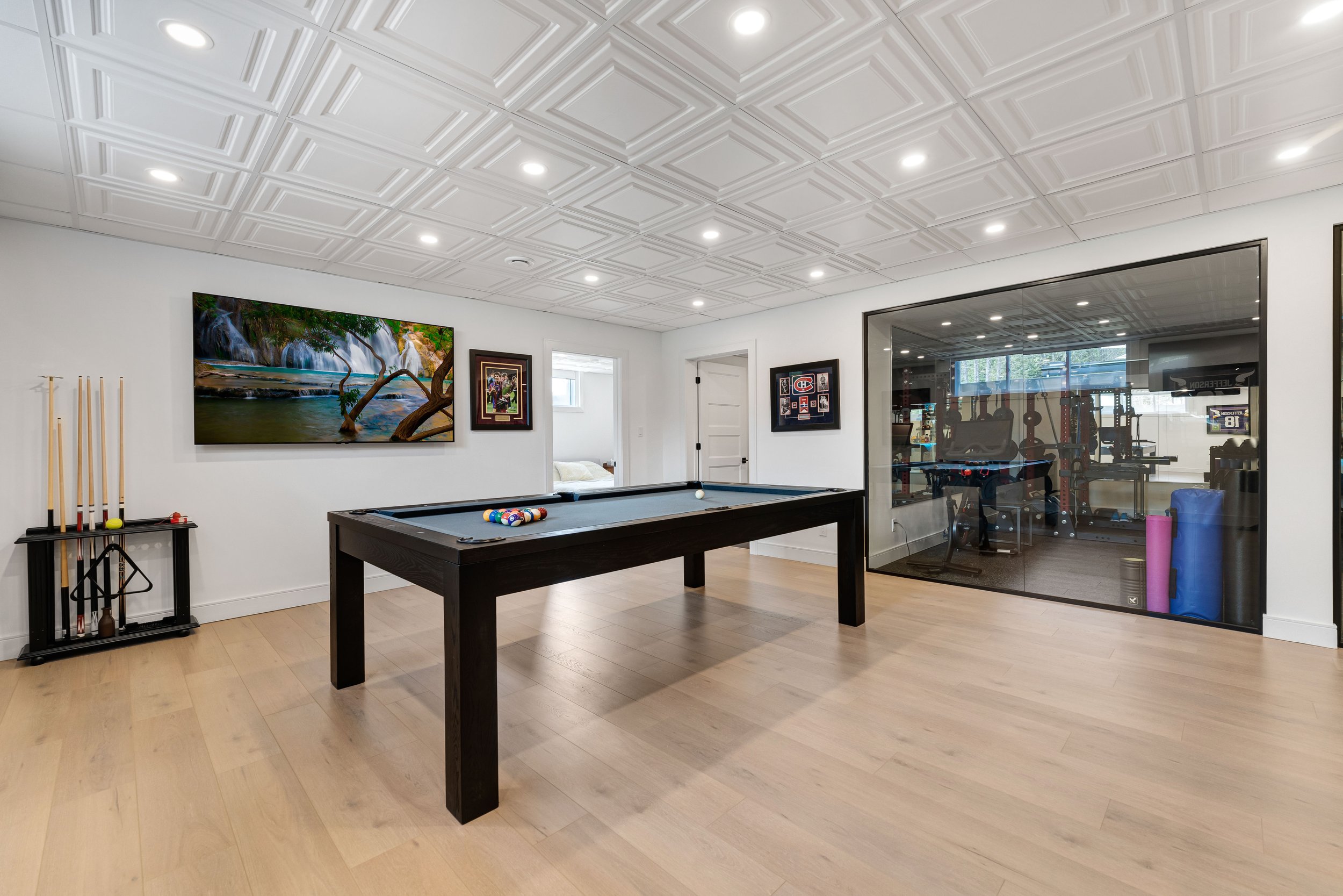 Modern basement recreation room with a pool table, exercise equipment behind glass wall, hardwood floor, and artwork on the walls.