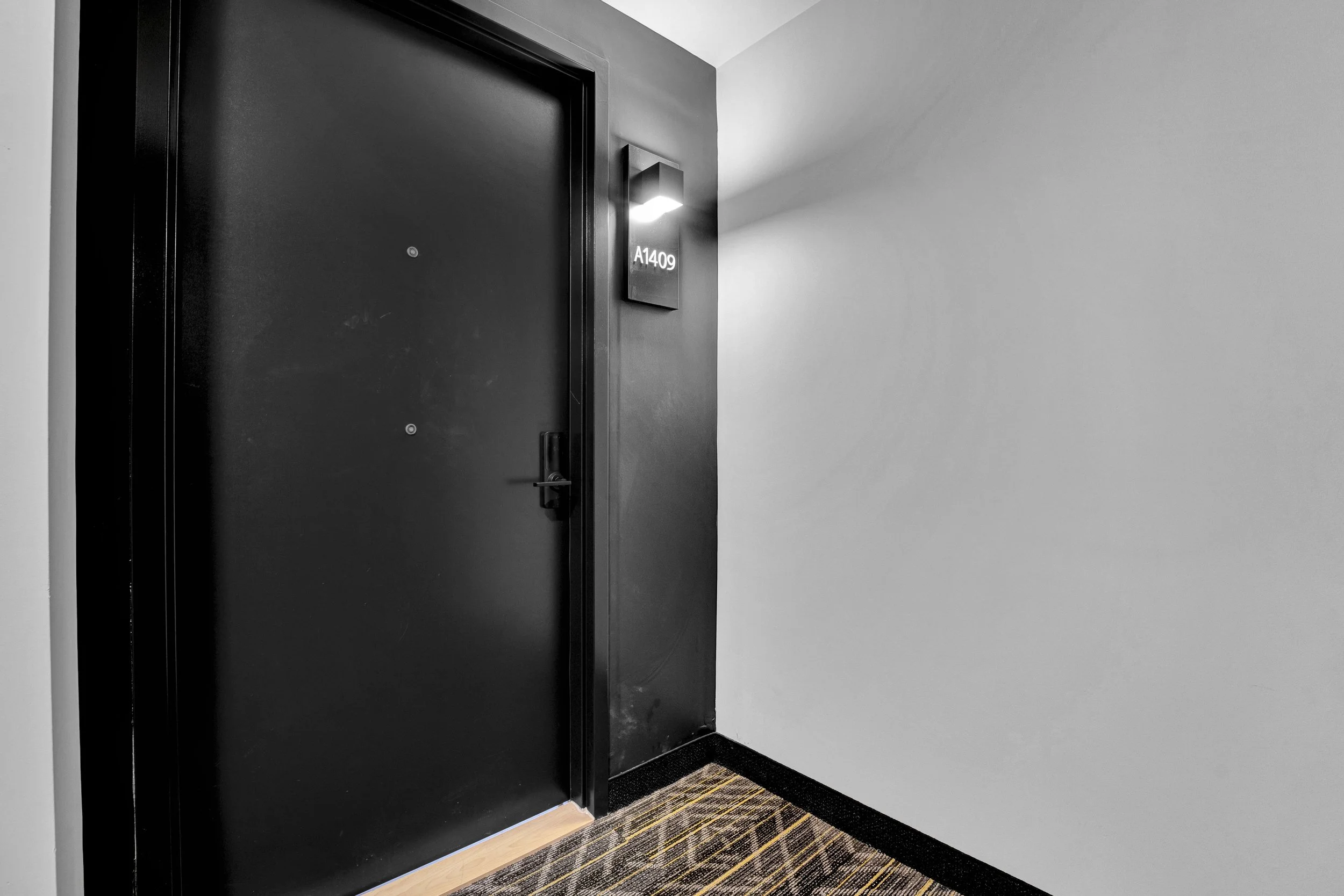 Black door with a peephole, metal handle, and an illuminated sign with the room number A1409 next to a wall-mounted light
