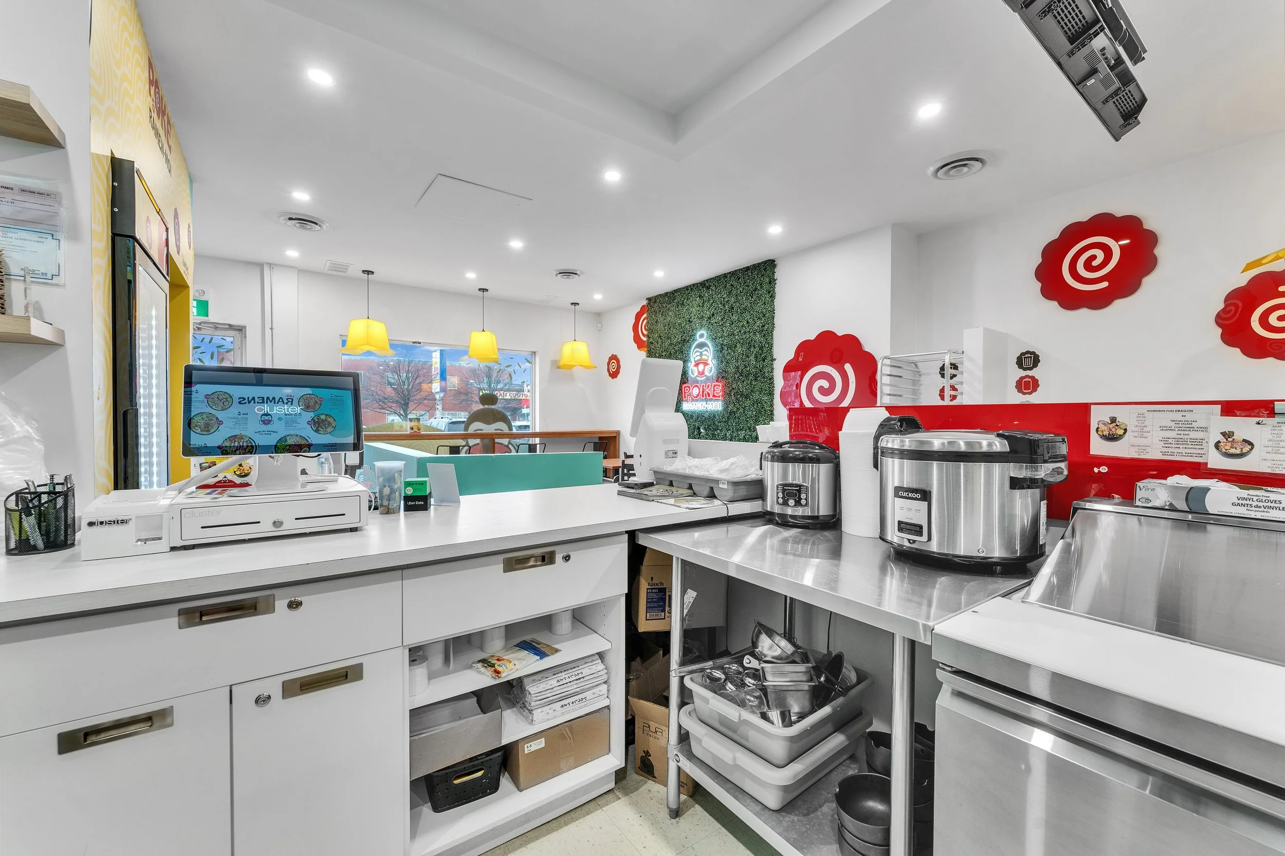 NVISION3D.ca Real Estate Media in Ottawa Interior of a fast food restaurant counter with a cash register, rice cooker, drink dispenser, and kitchen utensils, decorated with red swirl wall art and yellow hanging lights over seating area.