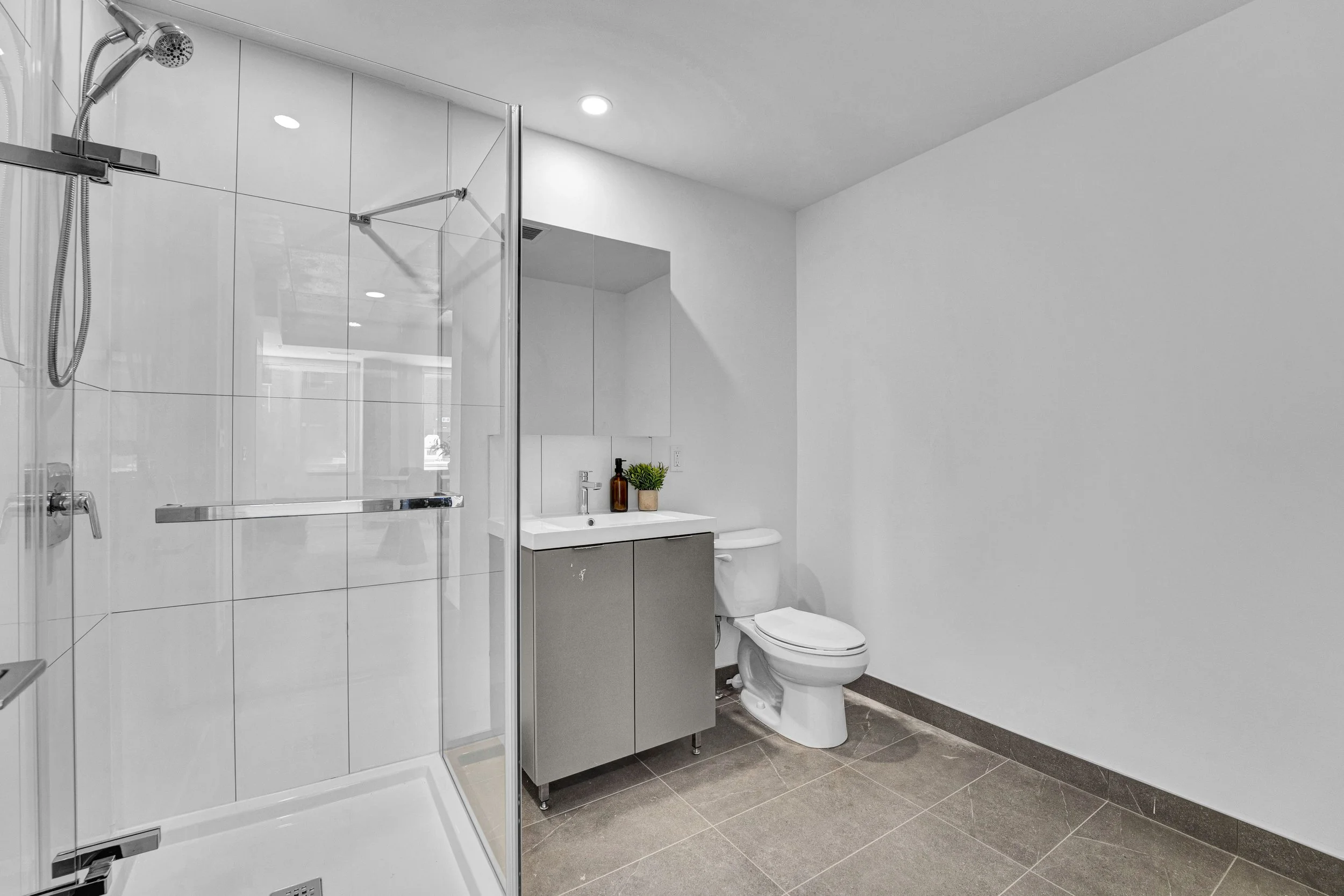 Ottawa Real Estate Media Services  Modern bathroom featuring a glass-enclosed shower, vanity with a sink, potted plants, and a toilet with a bidet attachment, all with white walls and tiled flooring.