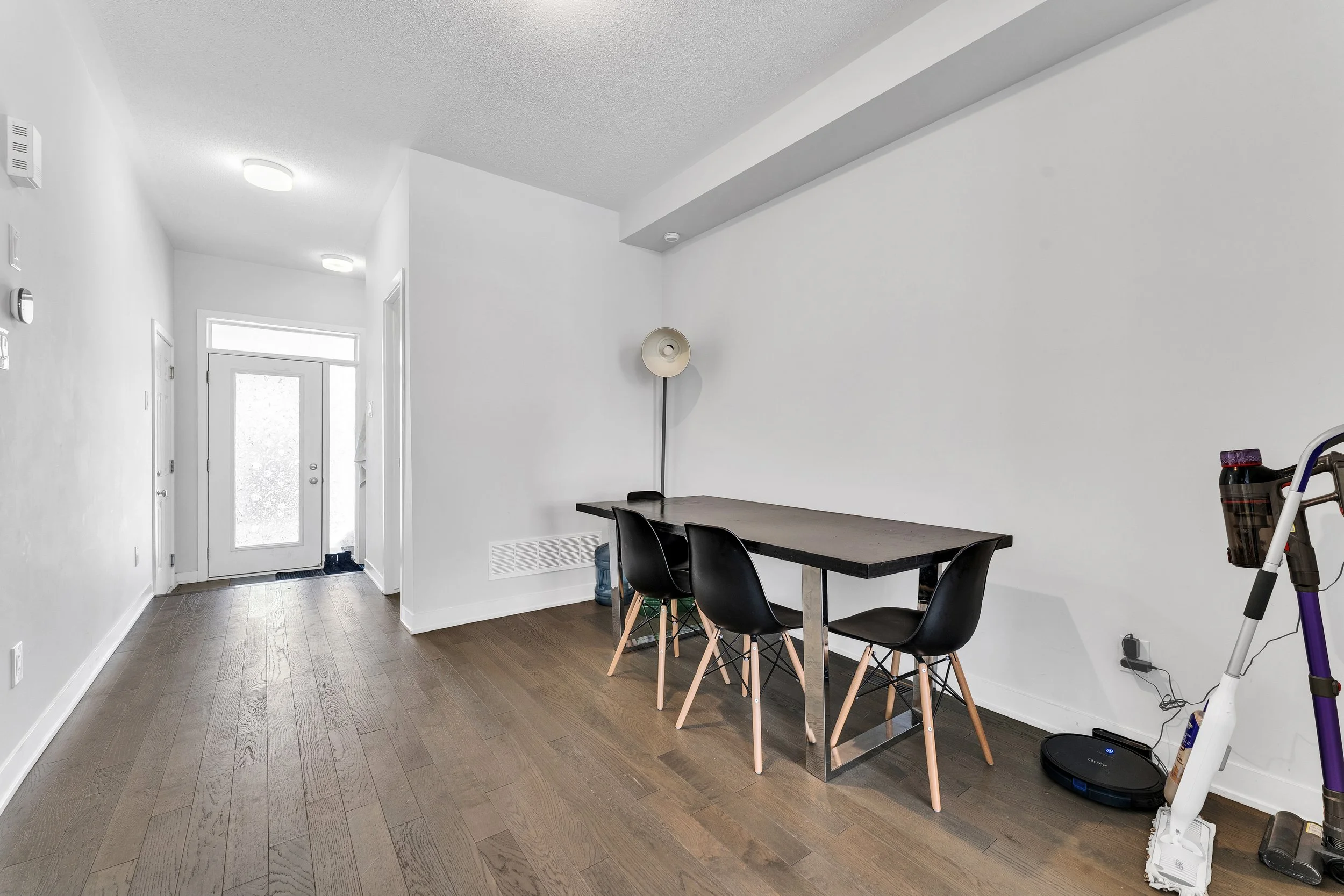 NV3D Real Estate PhotographInterior of a modern home entryway with a dining area, including a black table, three black chairs with wooden legs, a standing floor lamp, and a vacuum cleaner on hardwood flooring, with a white front door and white walls.