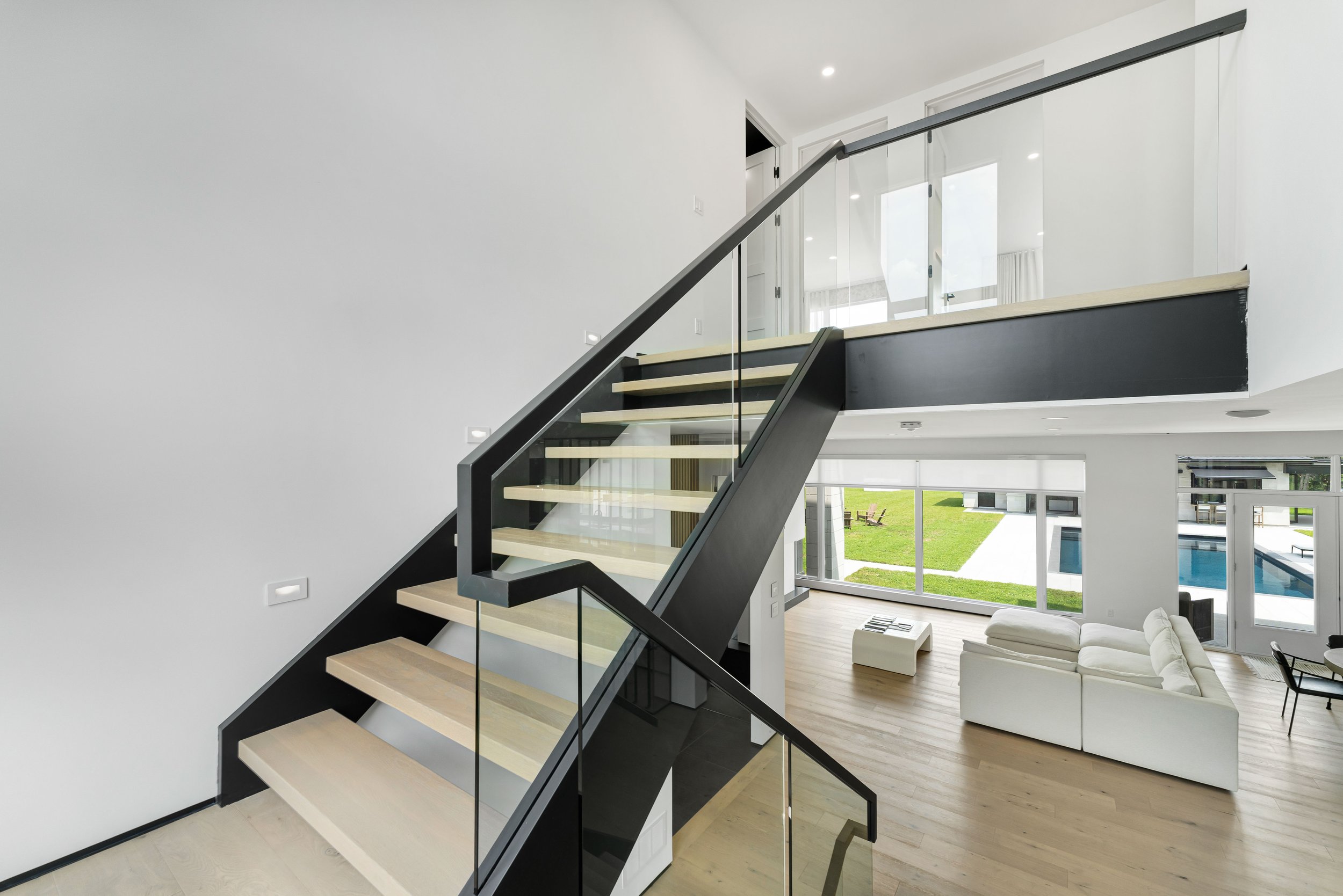 Modern interior with wooden staircase, glass railing, living room view with white sofa, coffee table, large windows, and garden with pool outside.