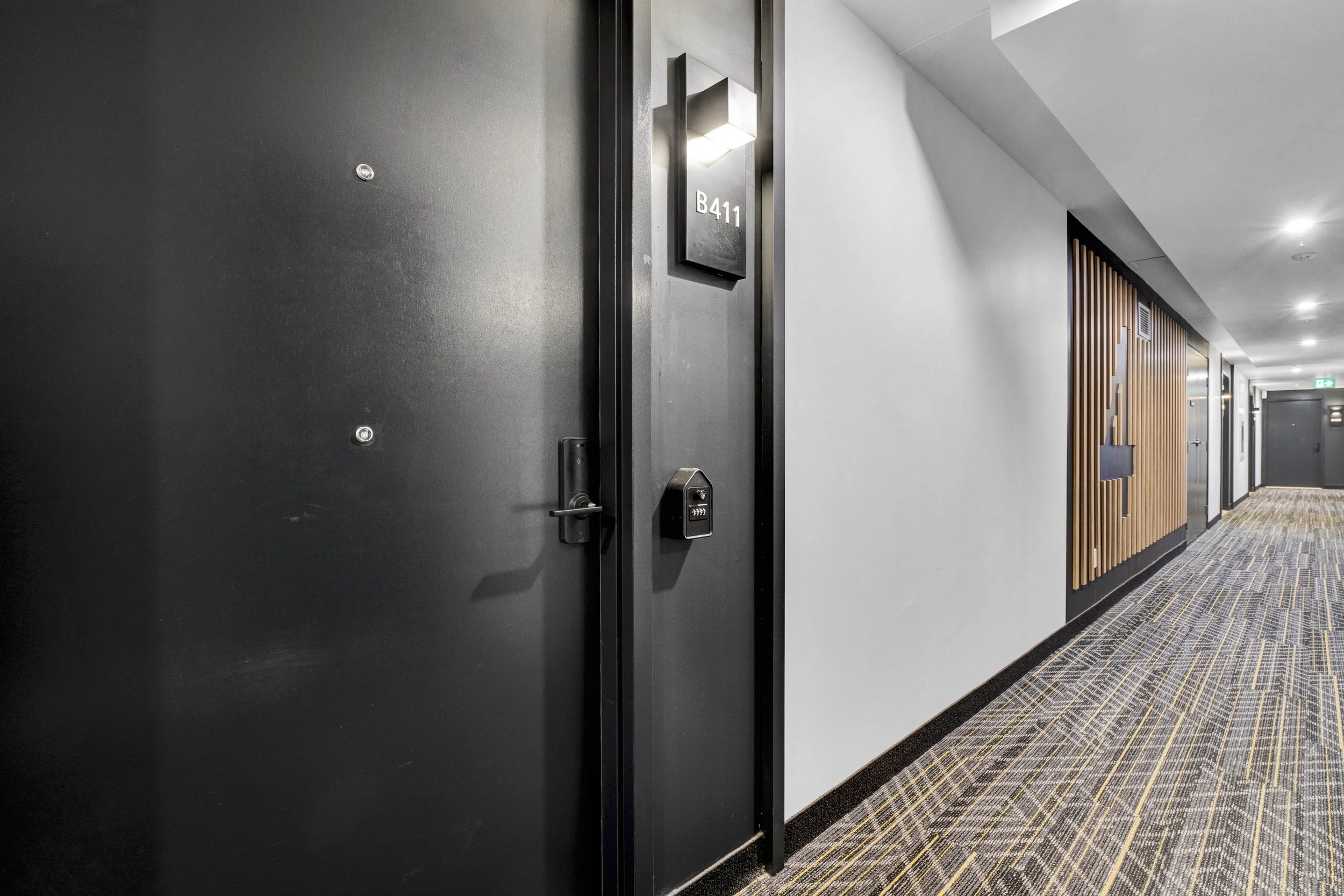 Ottawa Real Estate NVISION 3D  Lobby corridor of a hotel or office building with a black door labeled B411, a digital lock, and modern lighting. The hallway has a patterned carpet, white and wood-paneled walls, and ceiling lights.