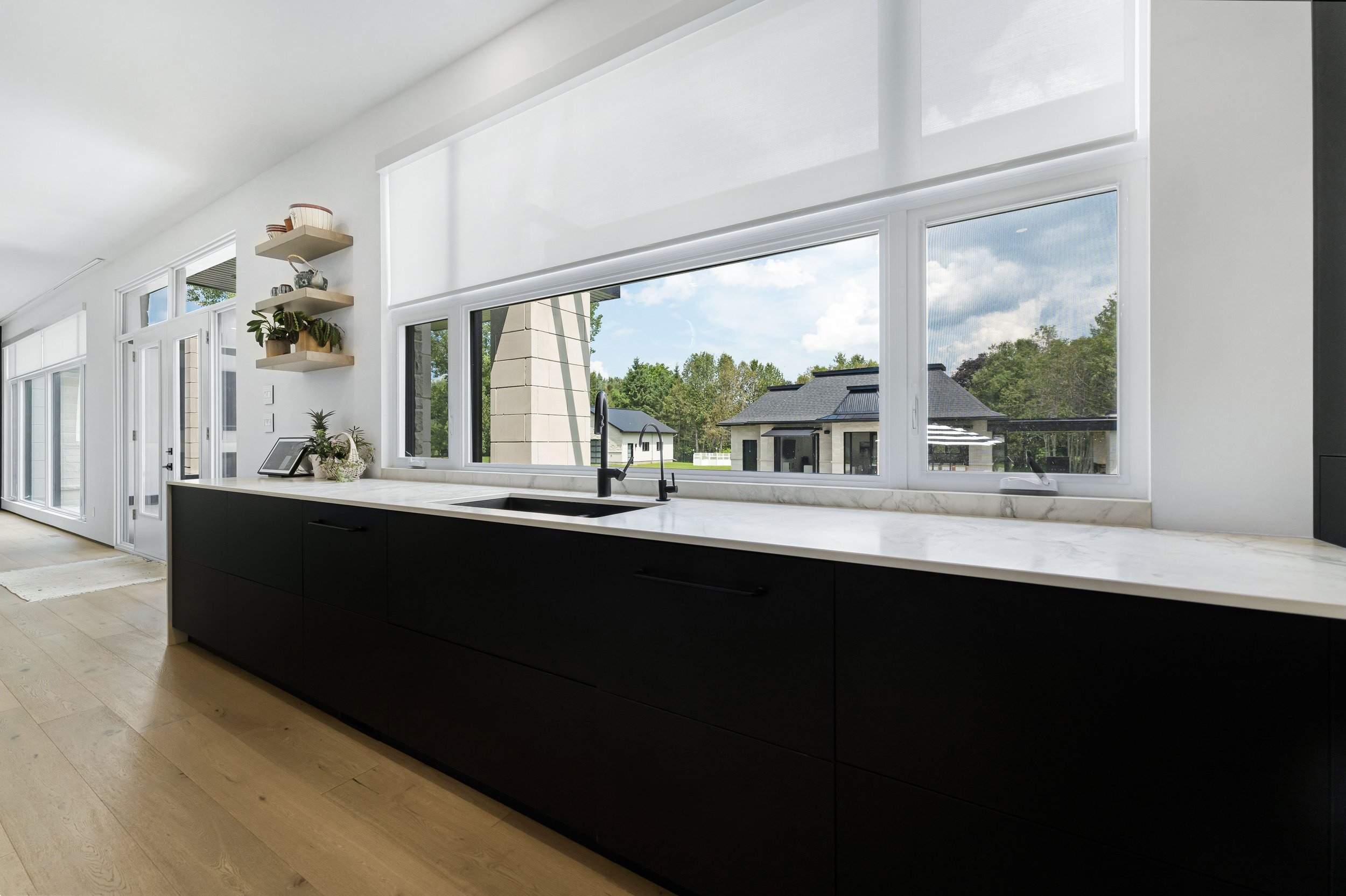 Modern kitchen with black cabinets, marble countertops, large windows, and outdoor view; wooden shelves with plants and decor.