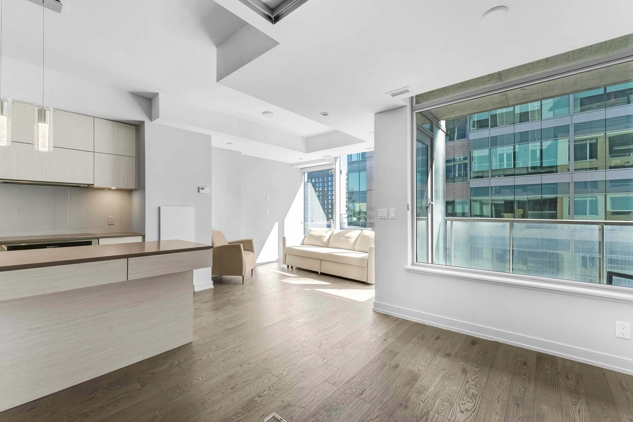 NV3D Ottawa Real Estate Photography Modern apartment living space with large windows, a white sofa, beige armchair, and a kitchen area with light wood cabinets.