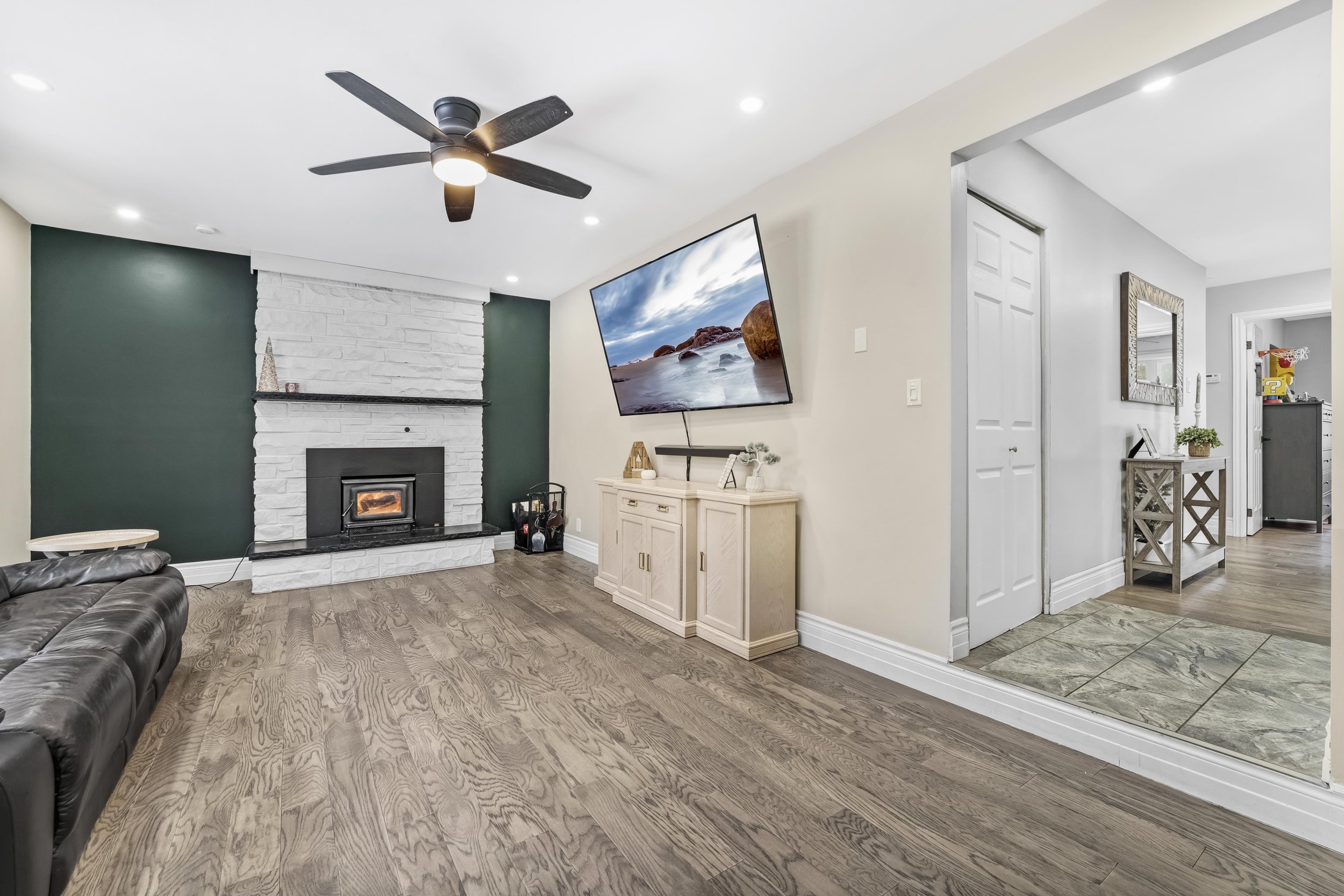 Real Estate Photography in Ottawa Living room with a white painted brick fireplace, green accent wall, wooden flooring, a ceiling fan, a TV mounted on the wall, and a doorway leading to another area of the house.