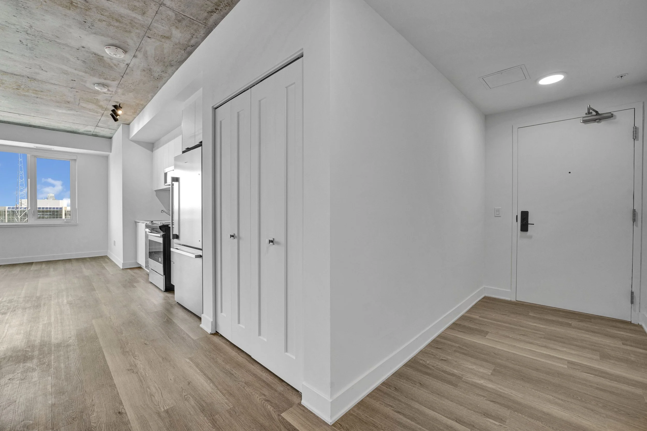 Interior view of a modern apartment with white walls, wooden flooring, and a ceiling with a concrete finish. The image shows a kitchen area with a window, kitchen appliances, and a white sliding closet door. There is a white door with a black handle,