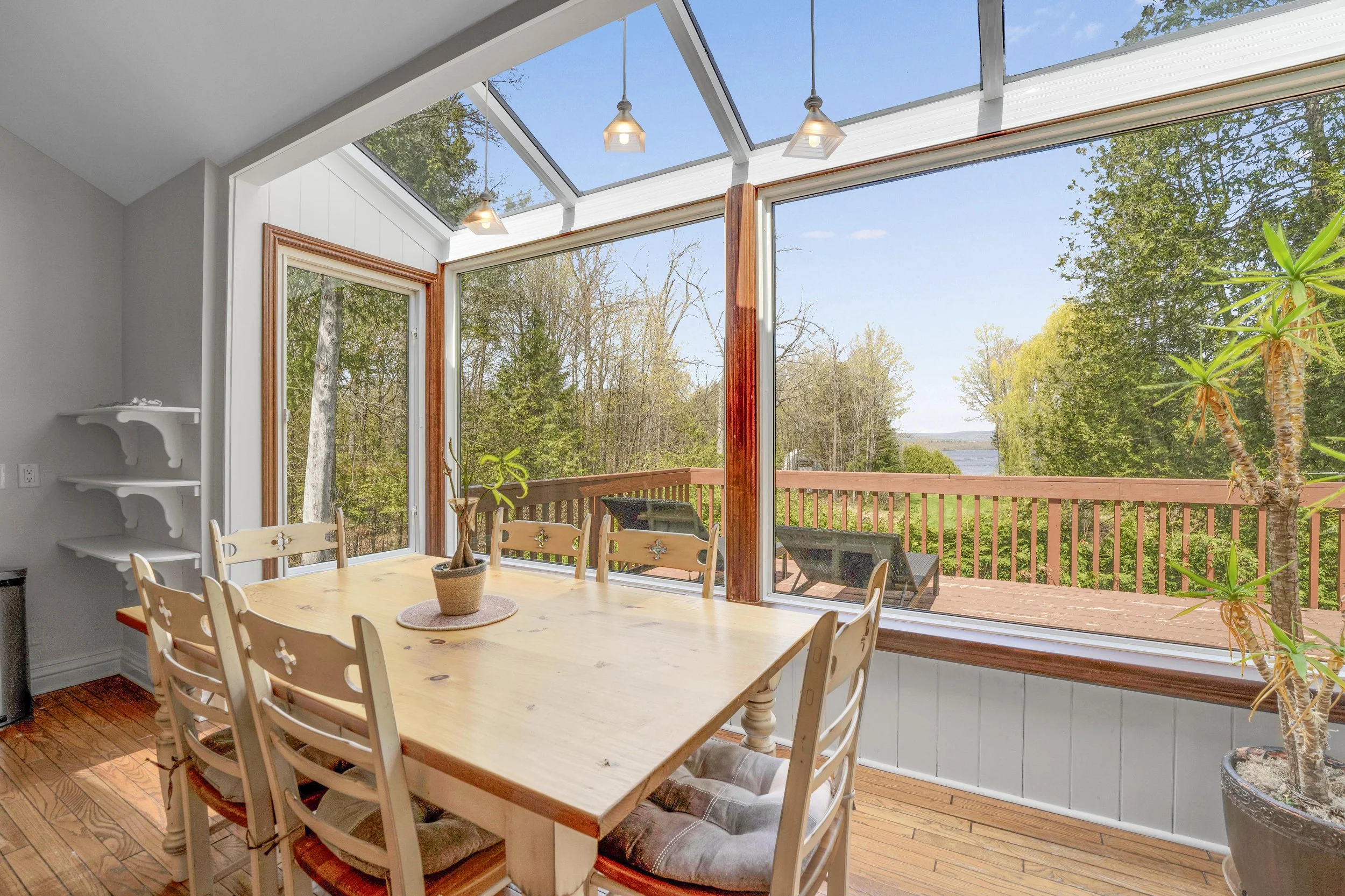 NVISION3D.ca Real Estate Media Ottawa  Sunroom with large glass windows and ceiling, wooden floor, a dining table, and chairs, overlooking trees and water outside.