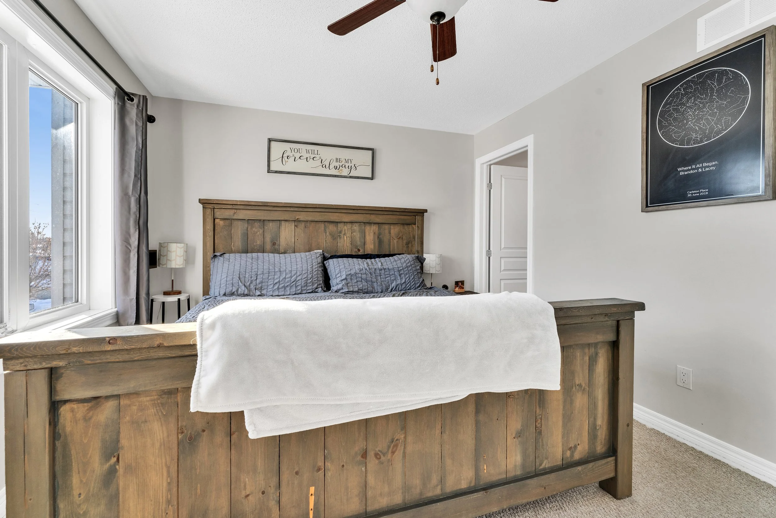A bedroom featuring a wooden bed with gray bedding, a window with curtains, a ceiling fan, and framed artwork on the walls.