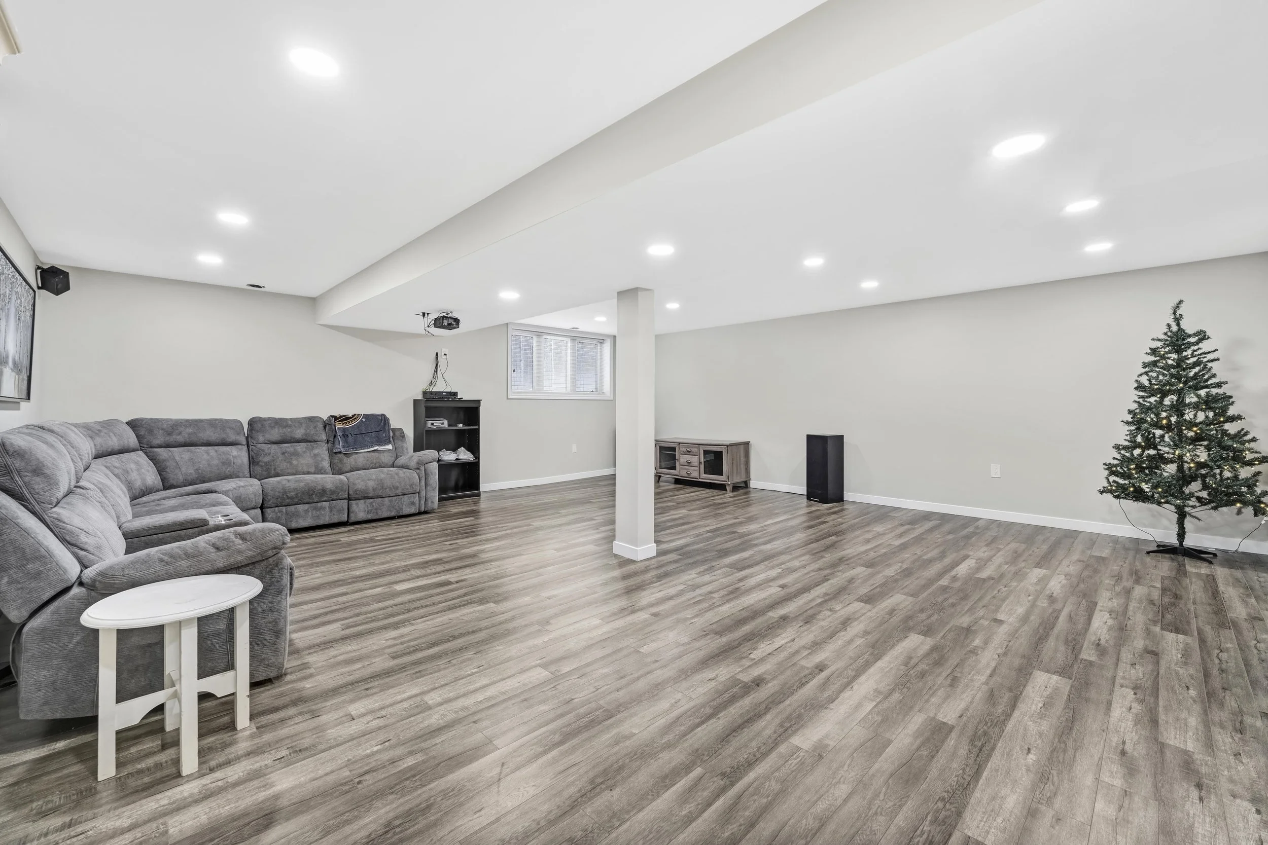 Ottawa Real Estate Photography  Spacious living room with gray sectional sofa, small white side table, and a Christmas tree, with recessed ceiling lights, wood laminate flooring, and a small window.