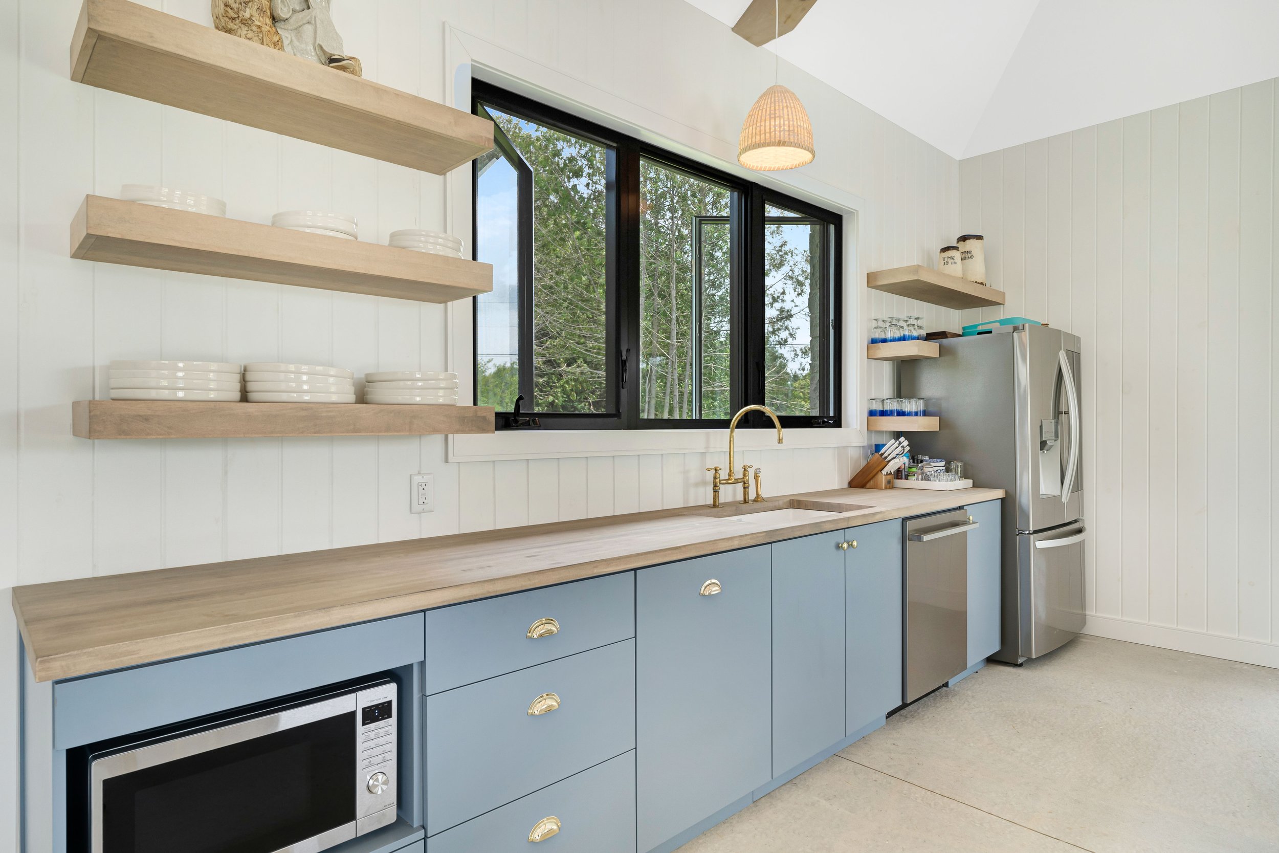 Modern kitchen interior with blue cabinets, stainless steel appliances, floating wooden shelves, dishes, and a large window.