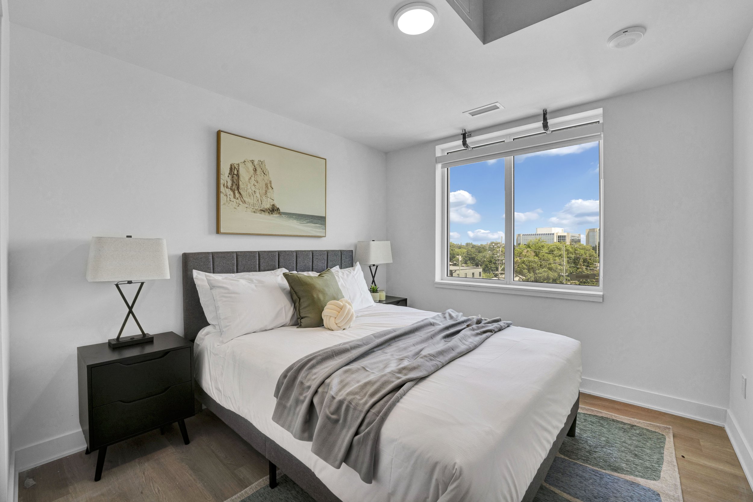 A modern bedroom with a large window showing a cityscape and blue sky. The bed has a gray headboard, white sheets, and pillows, with a gray blanket. There are two black bedside tables with white lamps, a framed picture of a beach on the wall, and a s