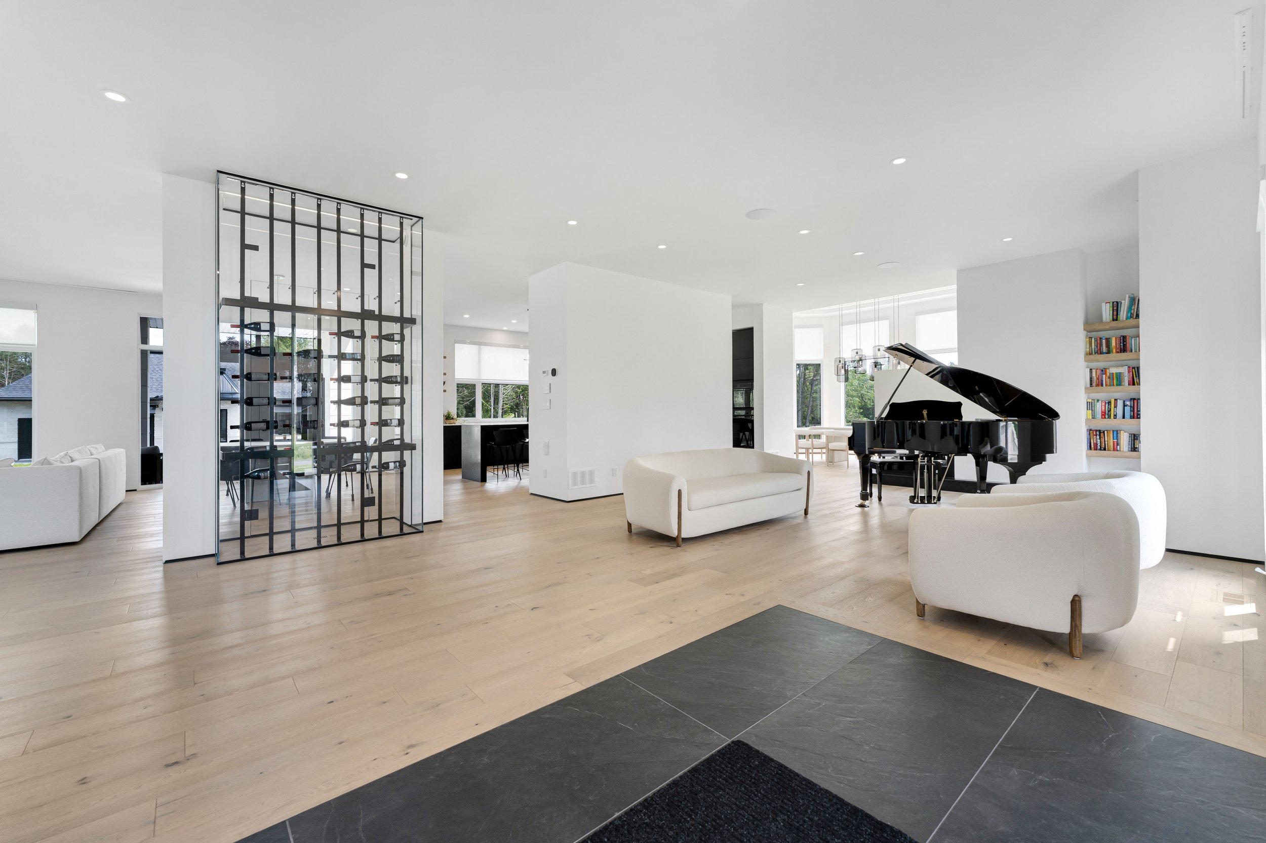 Modern open living space with white walls and light wood flooring. Features a black grand piano, white sofas, a bookshelf, and large windows. Stylish partition separates dining area with black furniture.