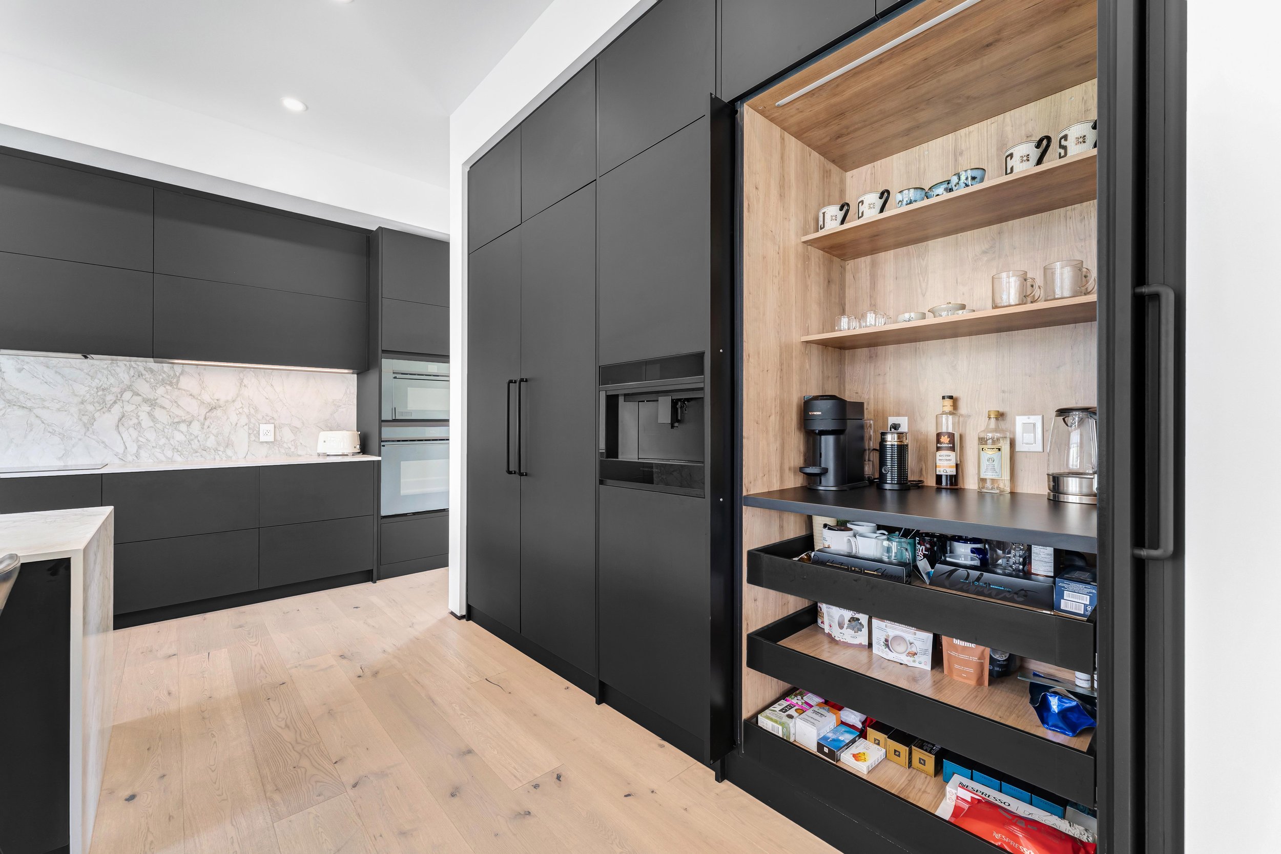 Modern kitchen with black cabinetry, marble backsplash, and light wood flooring. Open cabinet with coffee maker, mugs, and storage shelves visible.