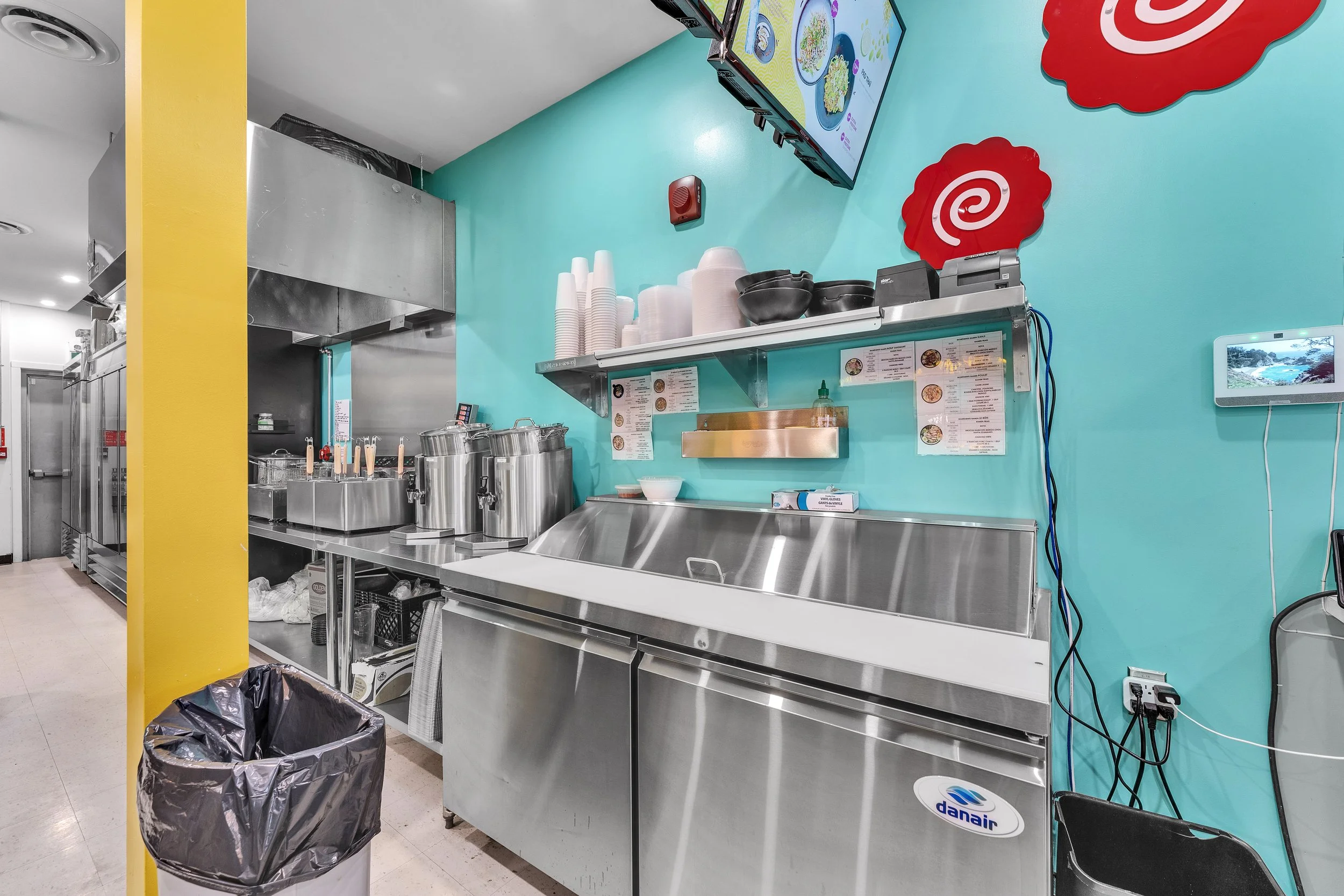 NVISION3D.ca Real Estate Media in Ottawa Interior of a restaurant kitchen with stainless steel counter and appliances, teal wall with red spiral logo decorations, menus, and supplies, including cups and bowls.