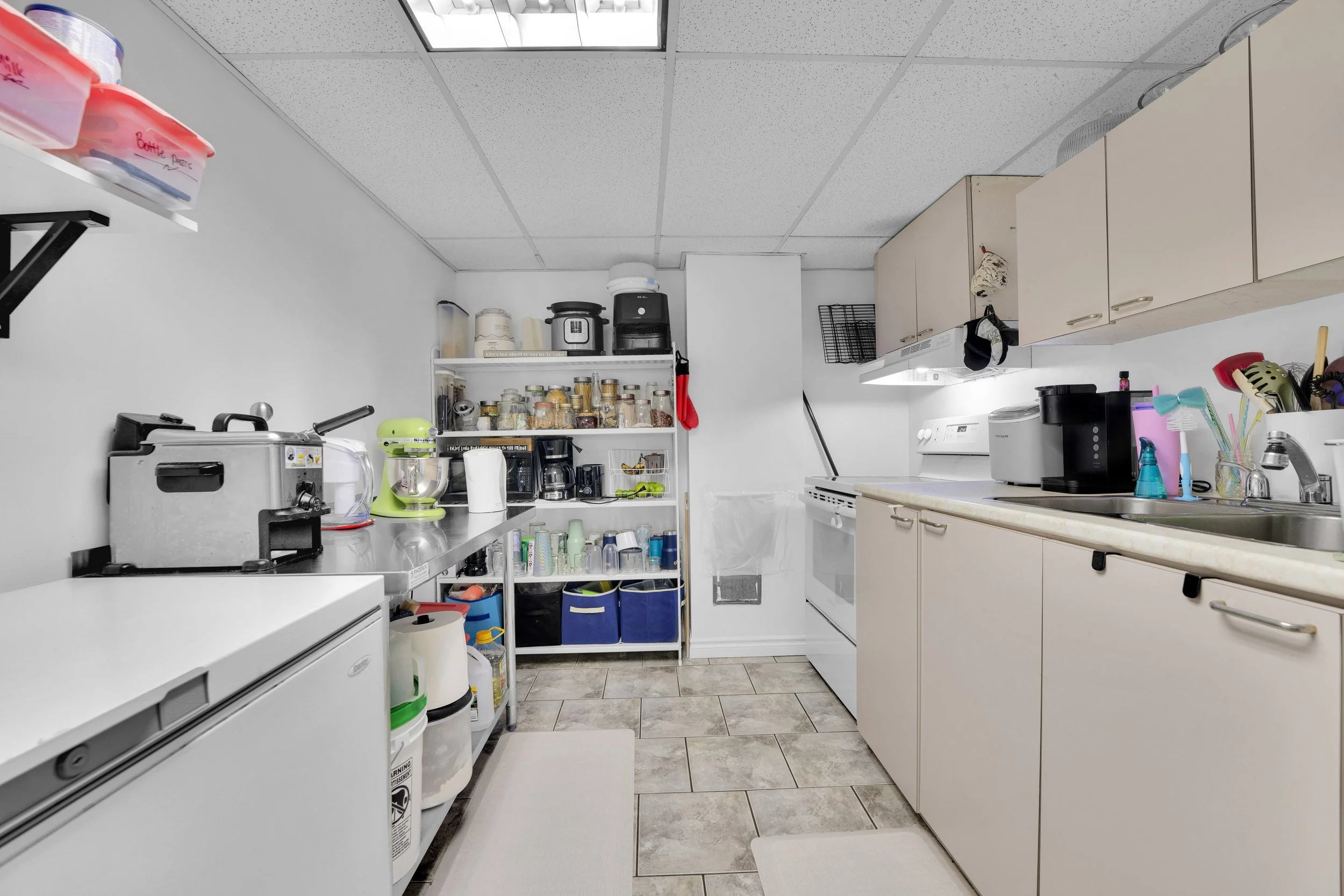Ottawa Real Estate Media Services  A small kitchen with white cabinets, tiled floor, and various appliances including a toaster oven, coffee maker, microwave, and sink. Shelves and counter space hold jars, bottles, utensils, and cleaning supplies.