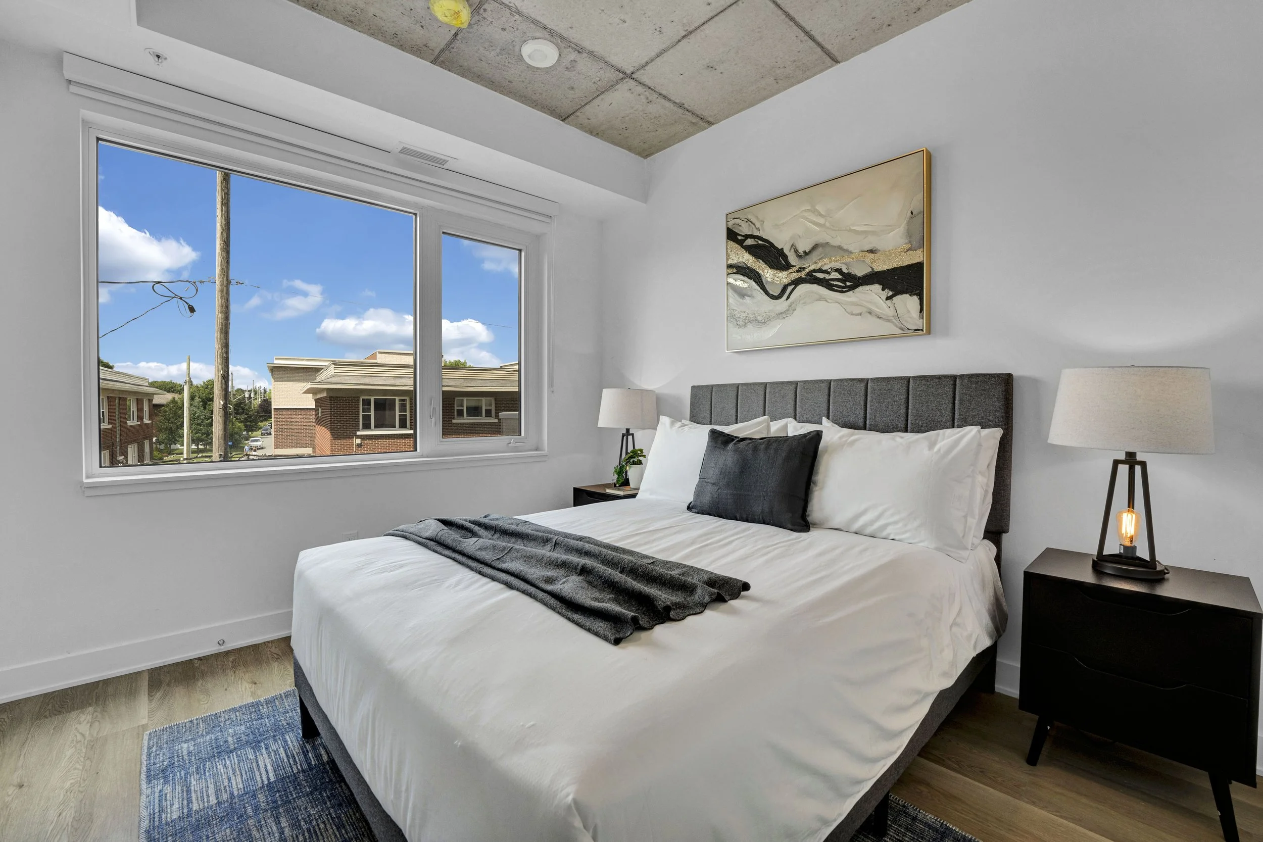Ottawa Real Estate NVISION 3D  A bedroom with a large window showing a clear blue sky, a bed with white sheets and dark pillows, a black nightstand with a lamp, and abstract wall art above the bed.