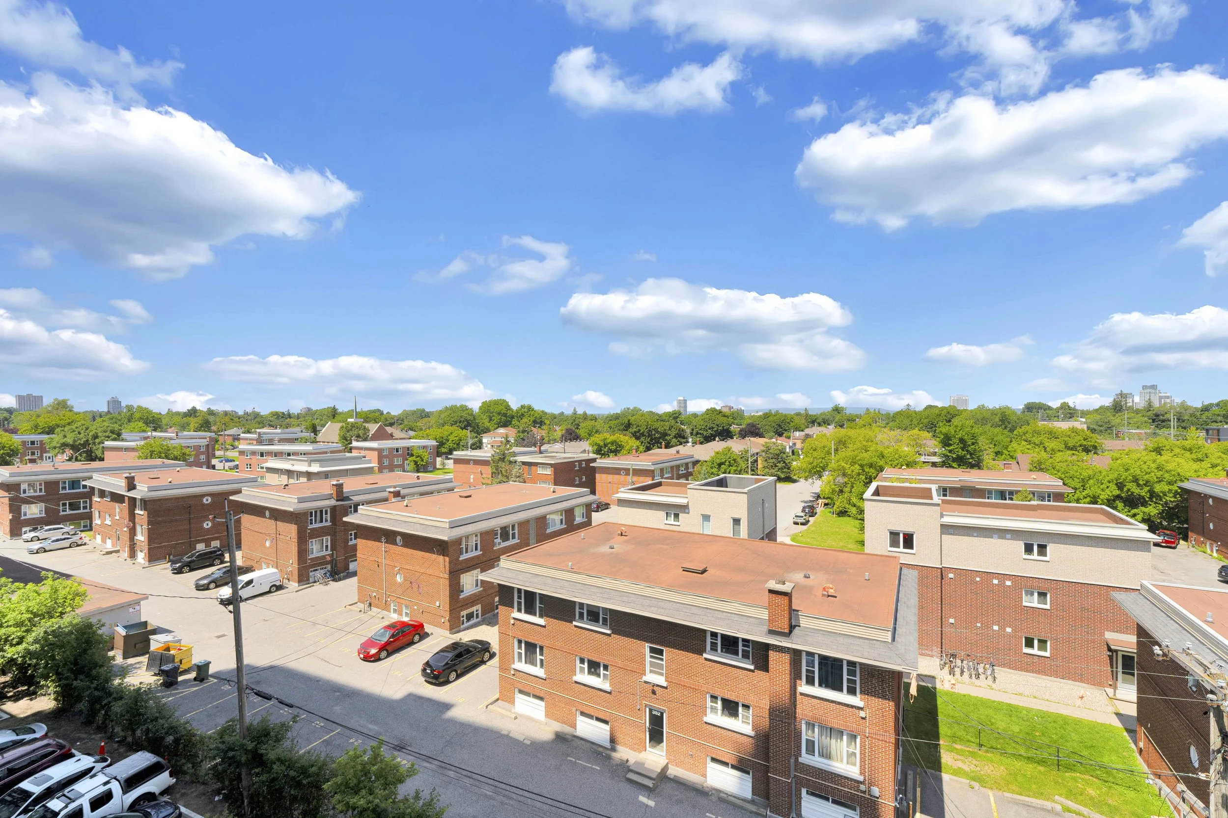 Ottawa Real Estate Media Services View of a neighborhood with multiple brick apartment buildings, parking lots with cars, green trees, and a blue sky with scattered clouds.