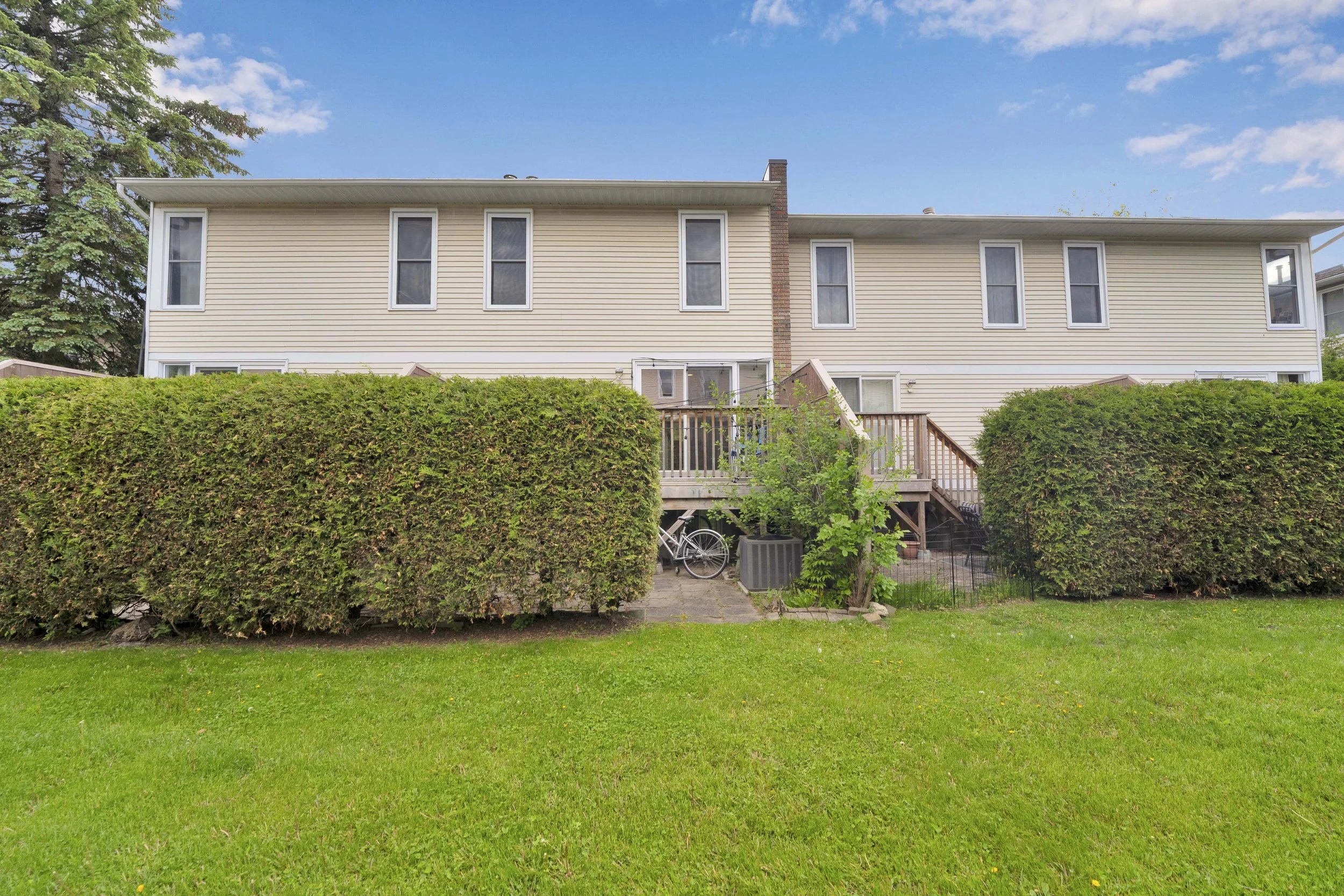 NVISION3D Real Estate Photography Ottawa  Backyard view of a beige two-story house with a wooden staircase leading to a door, surrounded by green bushes and a lush lawn, under a partly cloudy blue sky.
