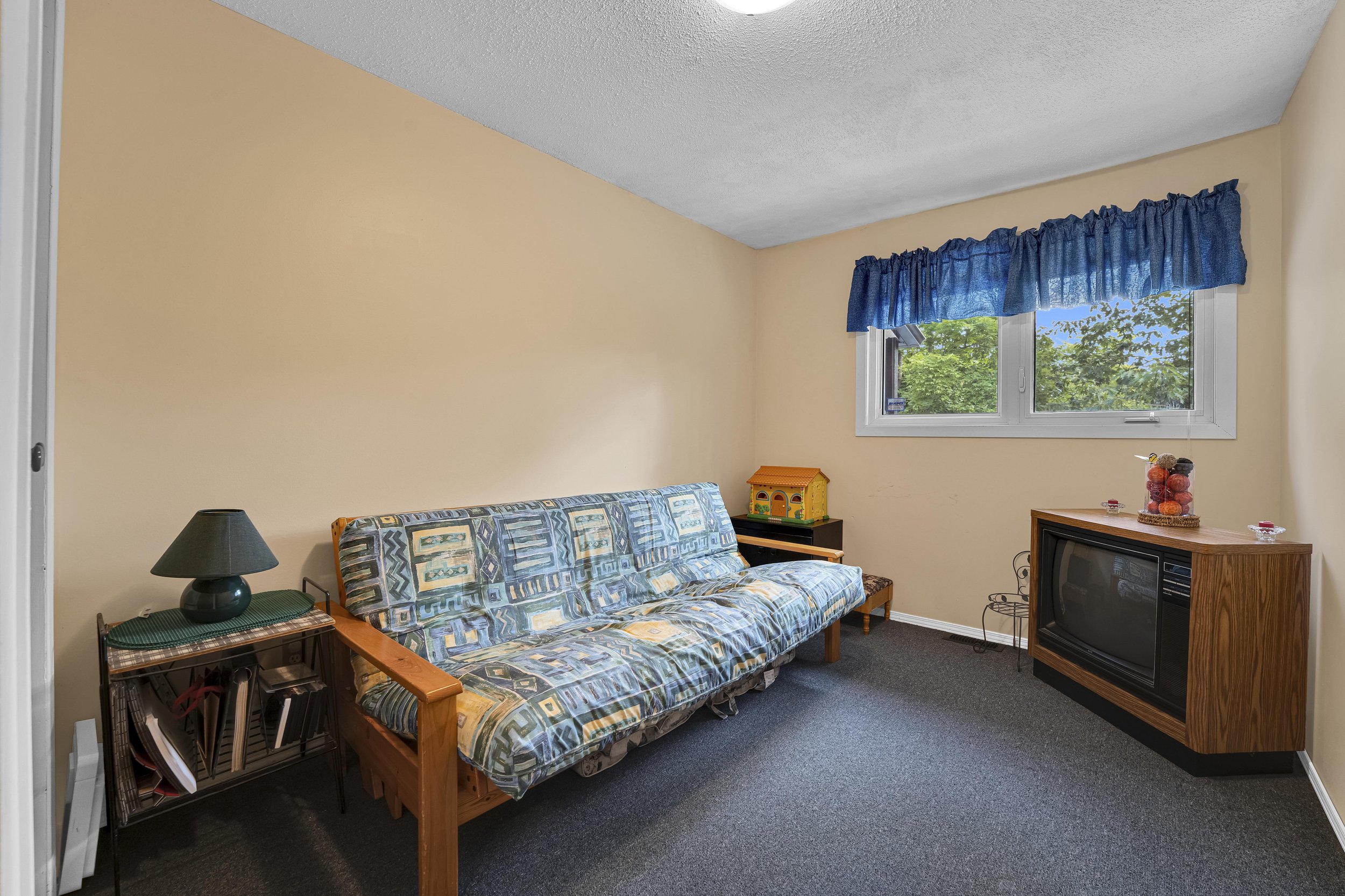 Ottawa Real Estate Photographer A cozy living room with a patterned couch, a small side table with a lamp, a wooden TV stand with a vintage television, a window with blue curtains, a toy house, and a vase with decorative balls.