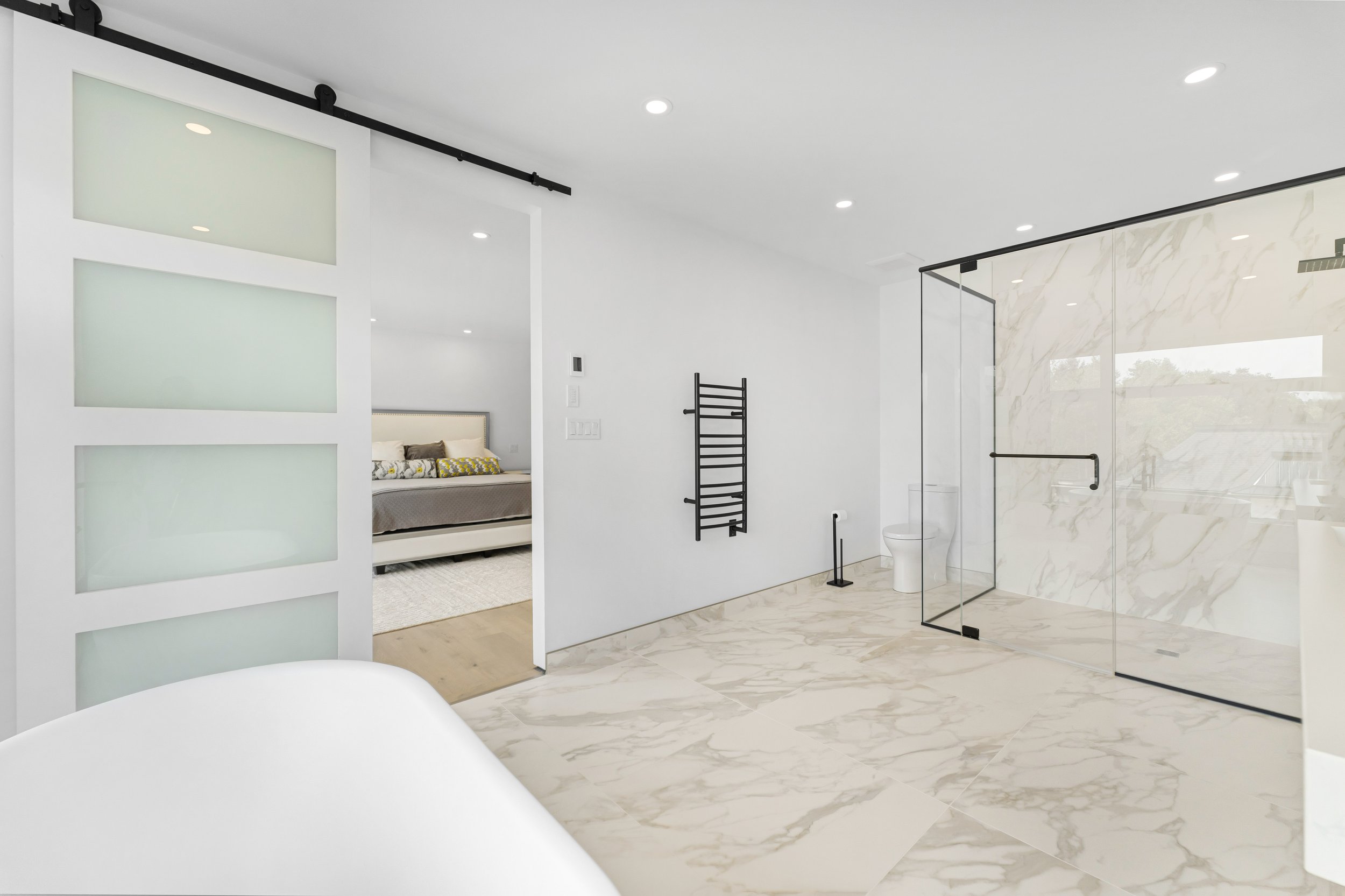 Modern bathroom with marble flooring, a sliding frosted glass door leading to a bedroom, a large glass-enclosed shower, freestanding bathtub, toilet, and a black towel rack.