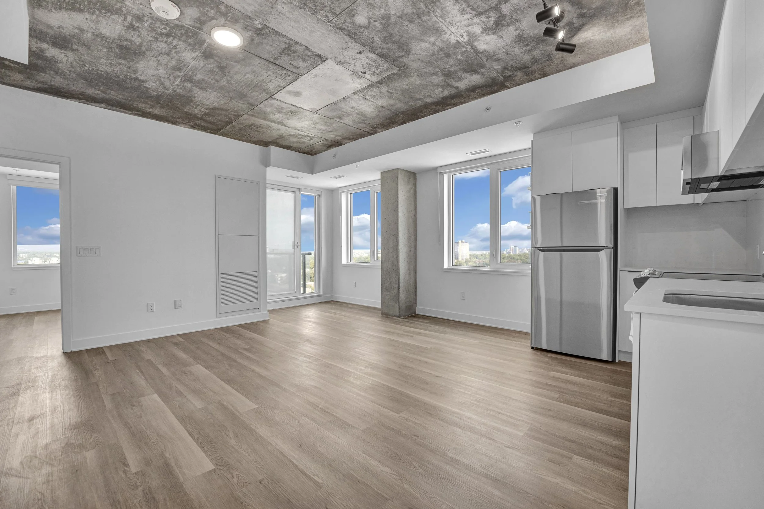 Interior of an unfurnished modern apartment with white walls, wood flooring, large windows showing a city view, a concrete ceiling, and a kitchen with white cabinets and a silver refrigerator.
