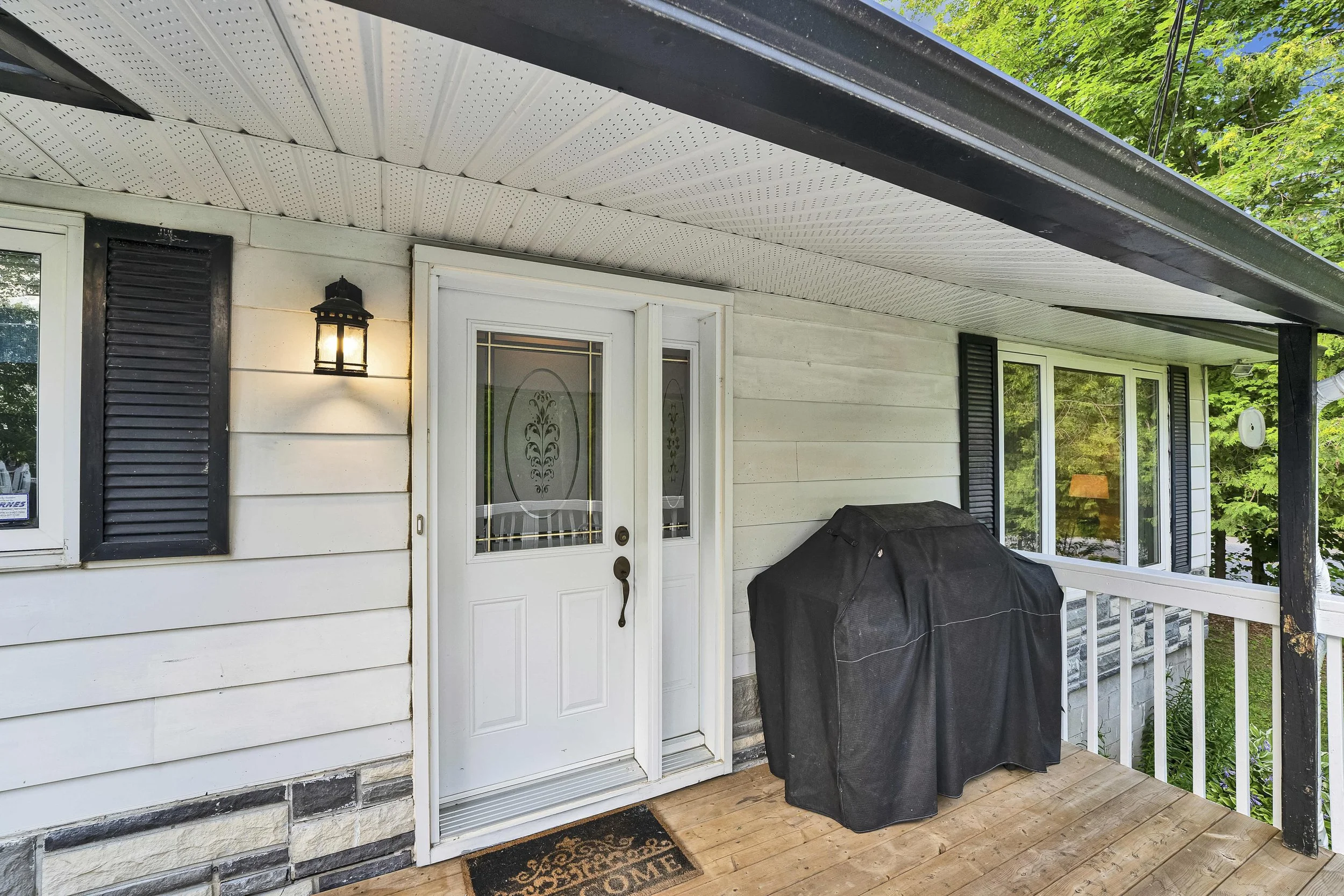 Ottawa Real Estate Photographer Back porch of a house with a white door, black shutters, outdoor light, a BBQ grill covered with a black cover, and a welcome mat on a wooden deck amid greenery.