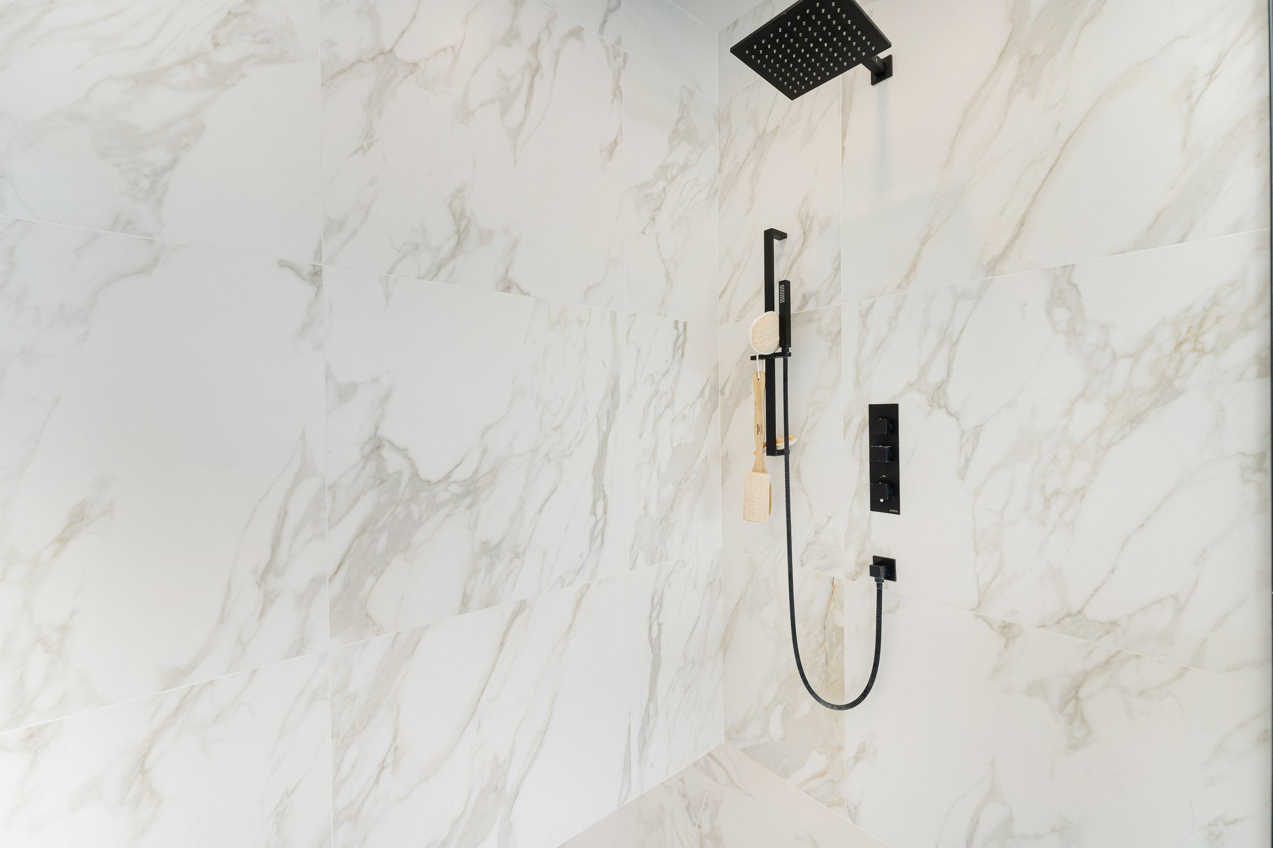 Modern shower with marble-patterned tiles, black rain shower head, and handheld shower attachment.