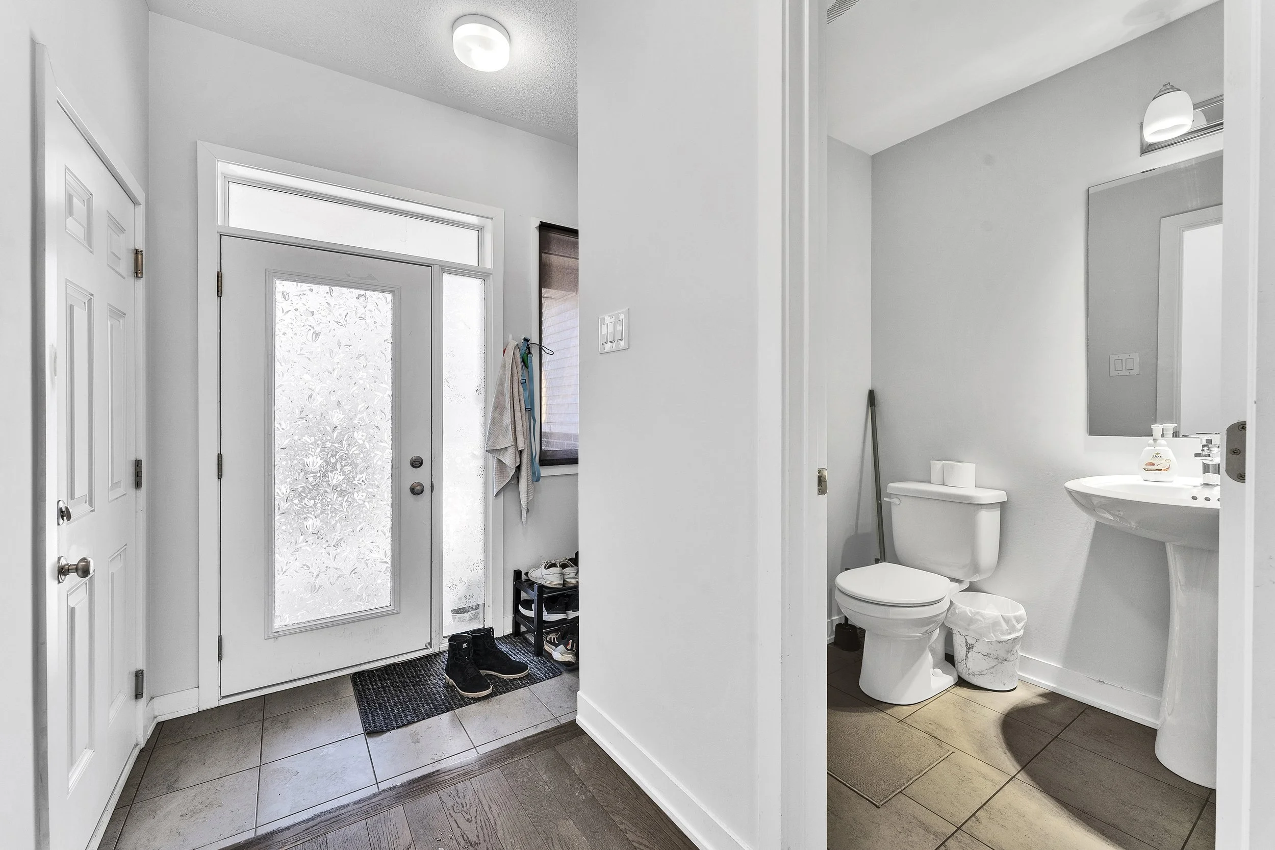 NV3D Real Estate Photography Ottawa  A small entryway with a white door and frosted glass window, shoes and a mat on the floor, and a view into a bathroom with a toilet, sink, mirror, and bathroom accessories.