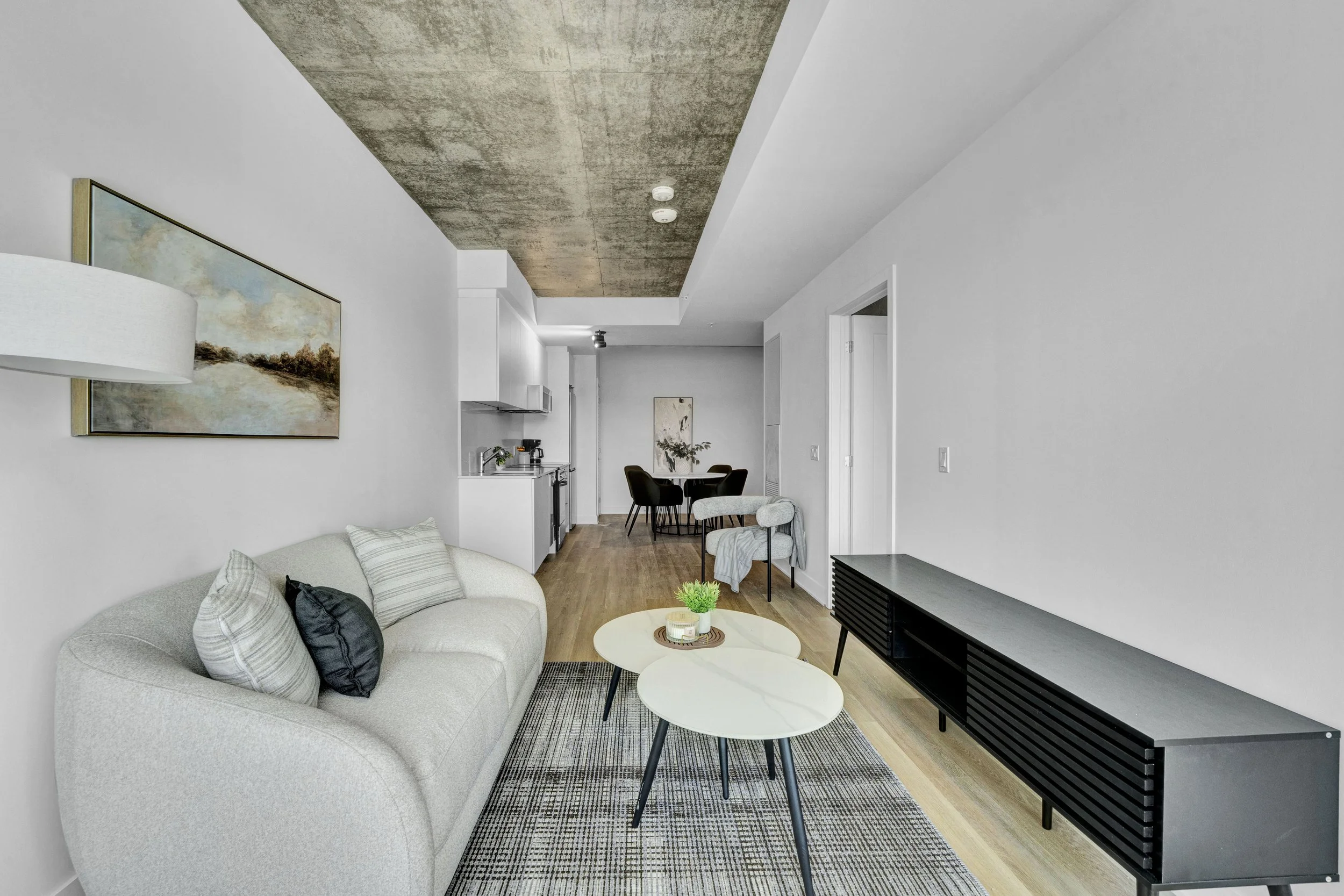 A modern living room with white walls, a light beige sofa with striped cushions, a black rug, and pair of round white coffee tables. There is a black TV console and an artwork hanging on the wall. In the background, a dining area with a round table a