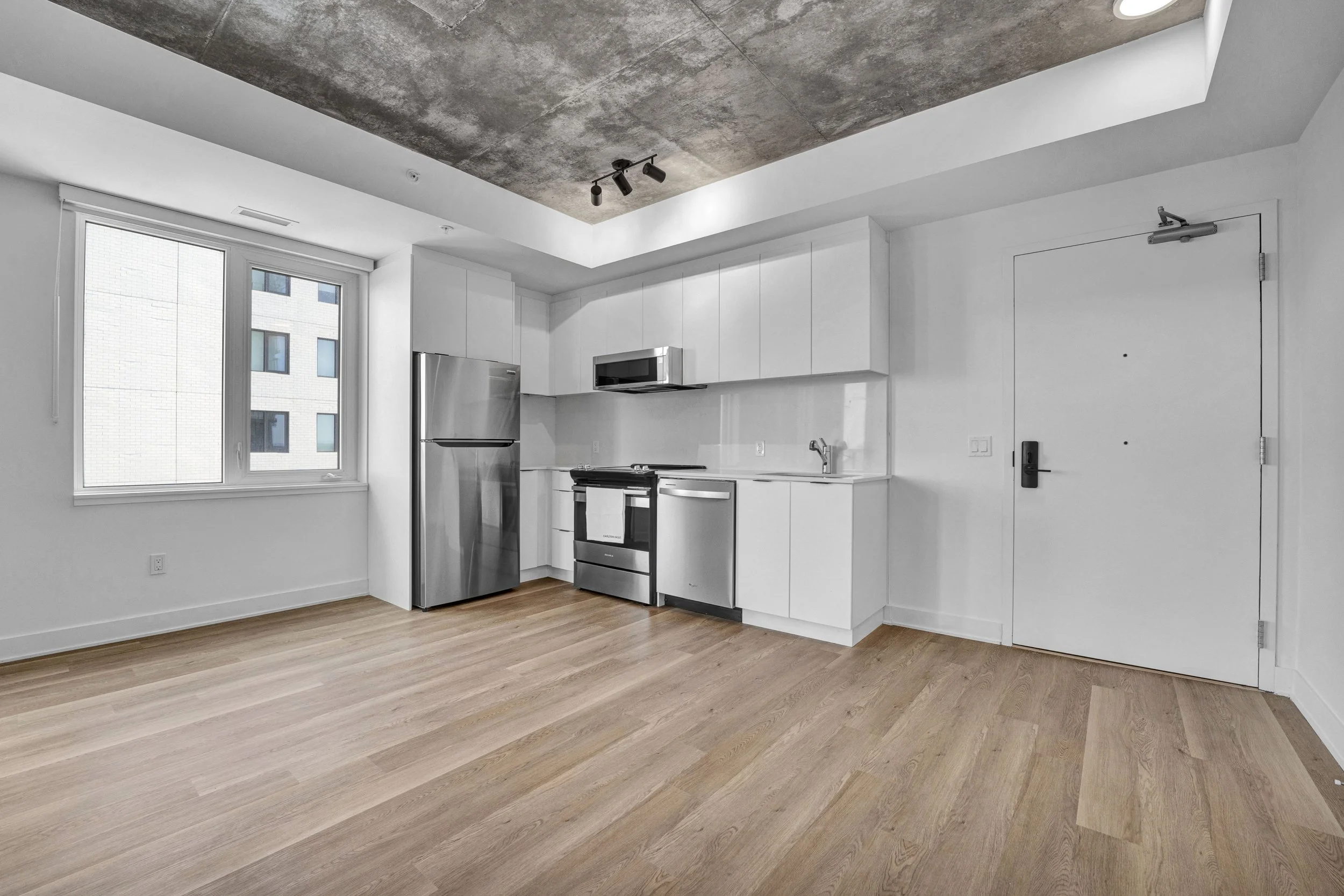 A minimalist kitchen with white cabinets, stainless steel appliances, a window, and a concrete ceiling.