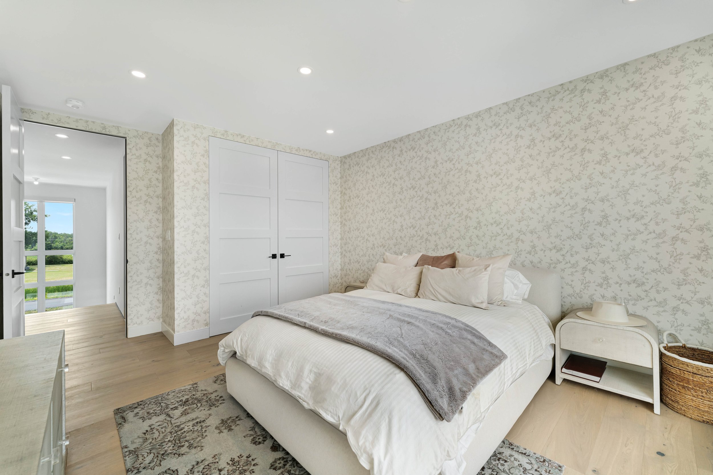 Bright bedroom with floral wallpaper, a neatly made bed with neutral bedding, a nightstand with a hat and book, a wicker basket, and white double closet doors.
