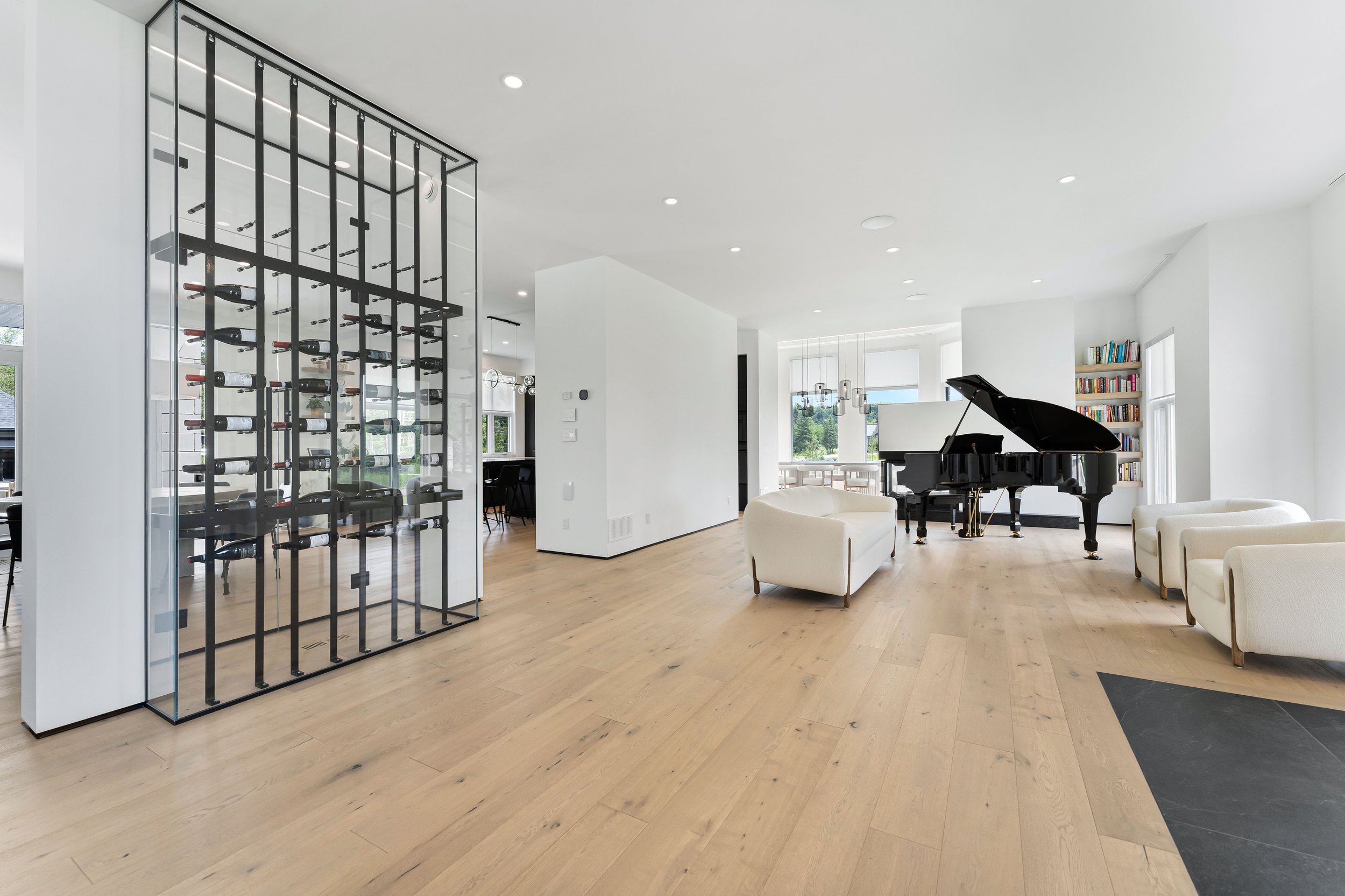 Modern living room with a glass wine rack, grand piano, white armchairs, hardwood floor, and bookshelves.