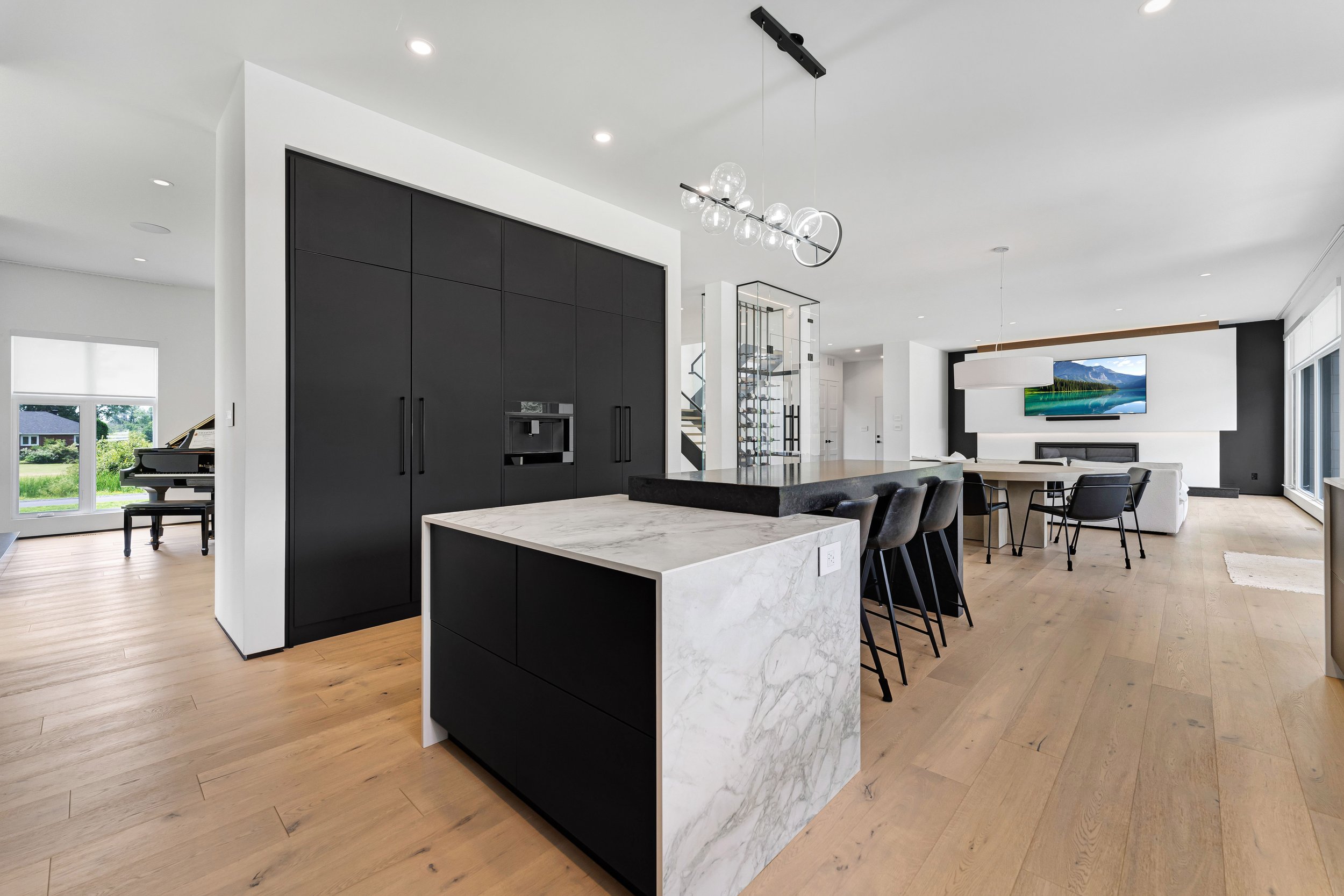 Modern open-plan kitchen and living area with hardwood flooring, black cabinets, marble countertops, dining table with chairs, large windows, and a grand piano in the background.