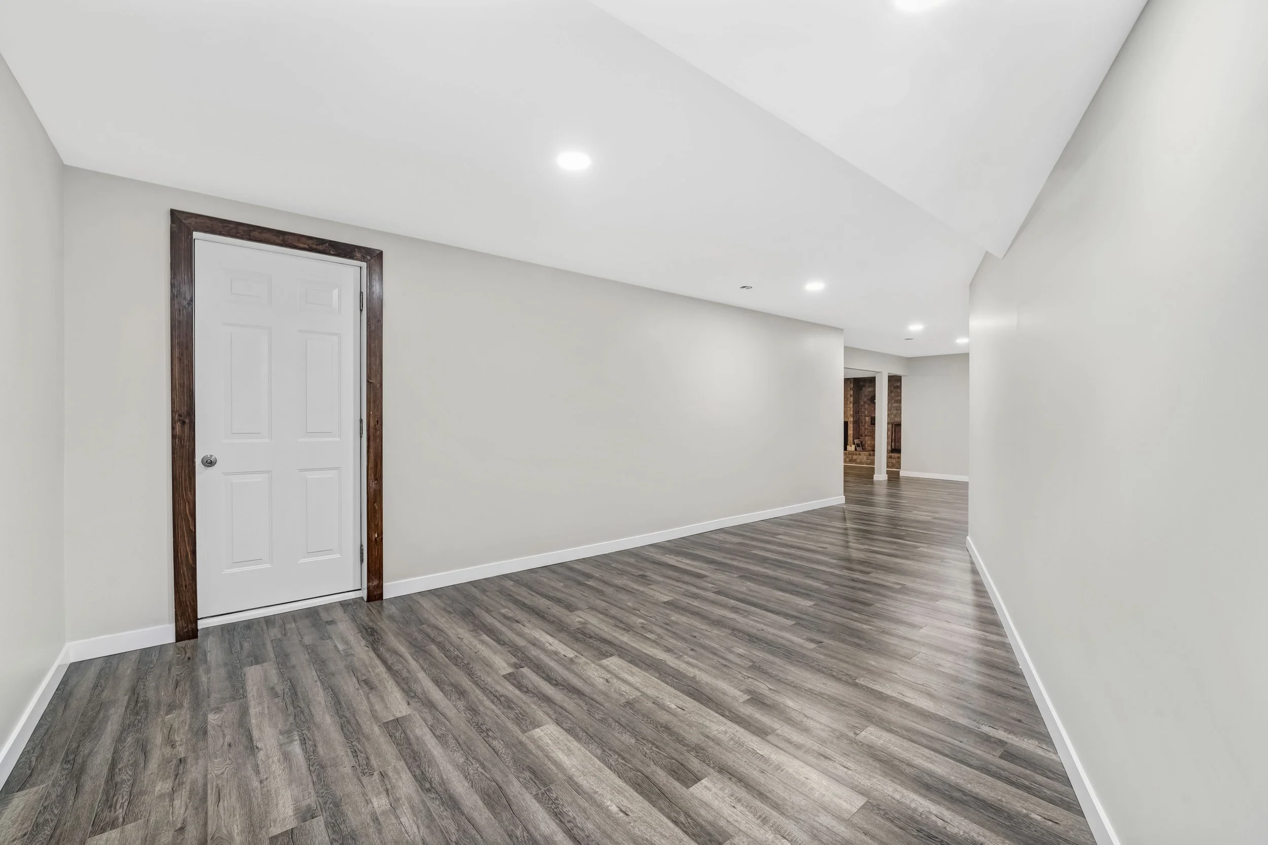 Empty living space with white walls, hardwood flooring, a white door with wooden trim, and recessed ceiling lights.