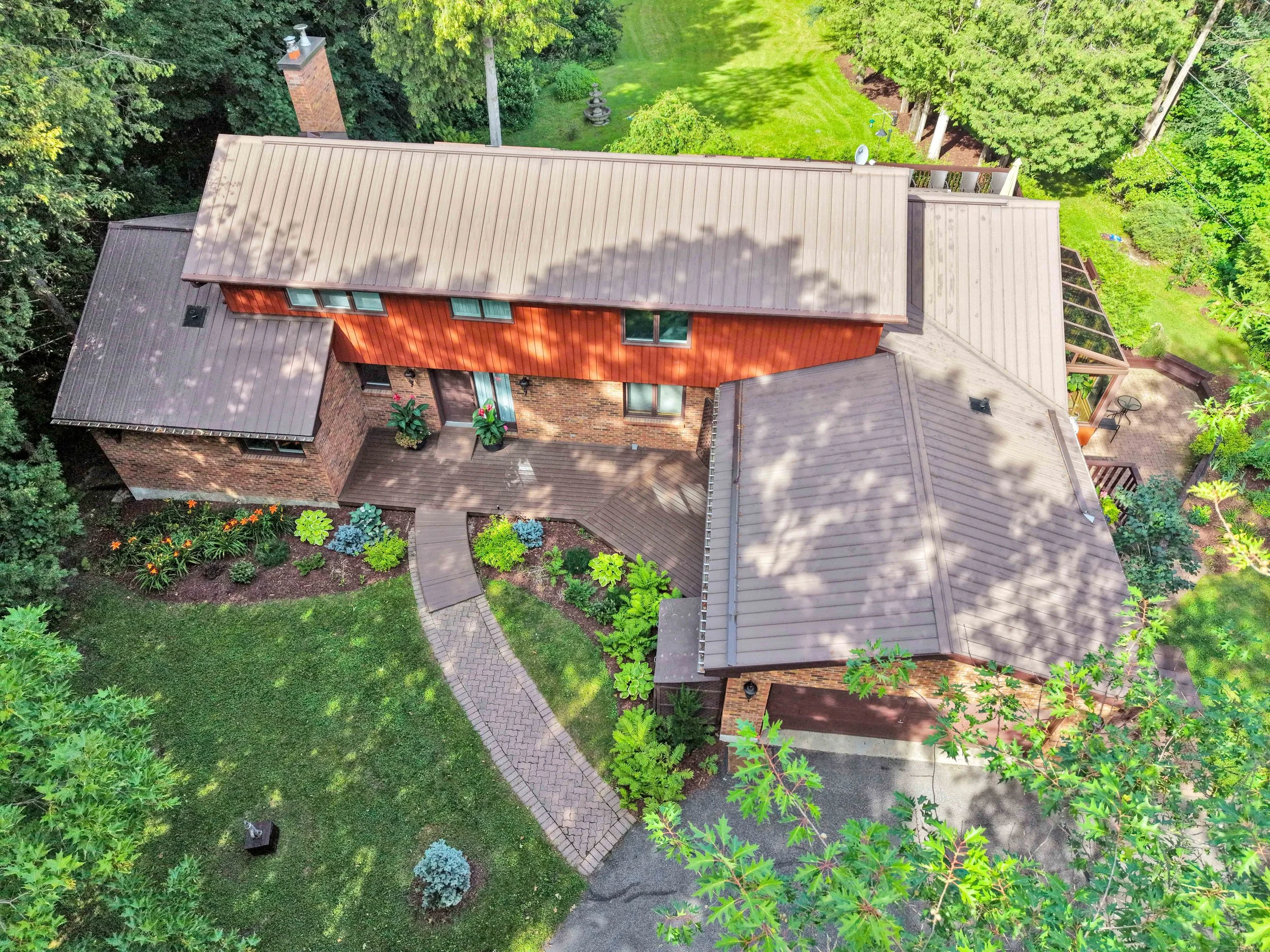 NVISION 3D Real Estate Media  Aerial view of a house with a brown roof, surrounded by lush green trees and a well-maintained garden with colorful plants and a paved walkway.