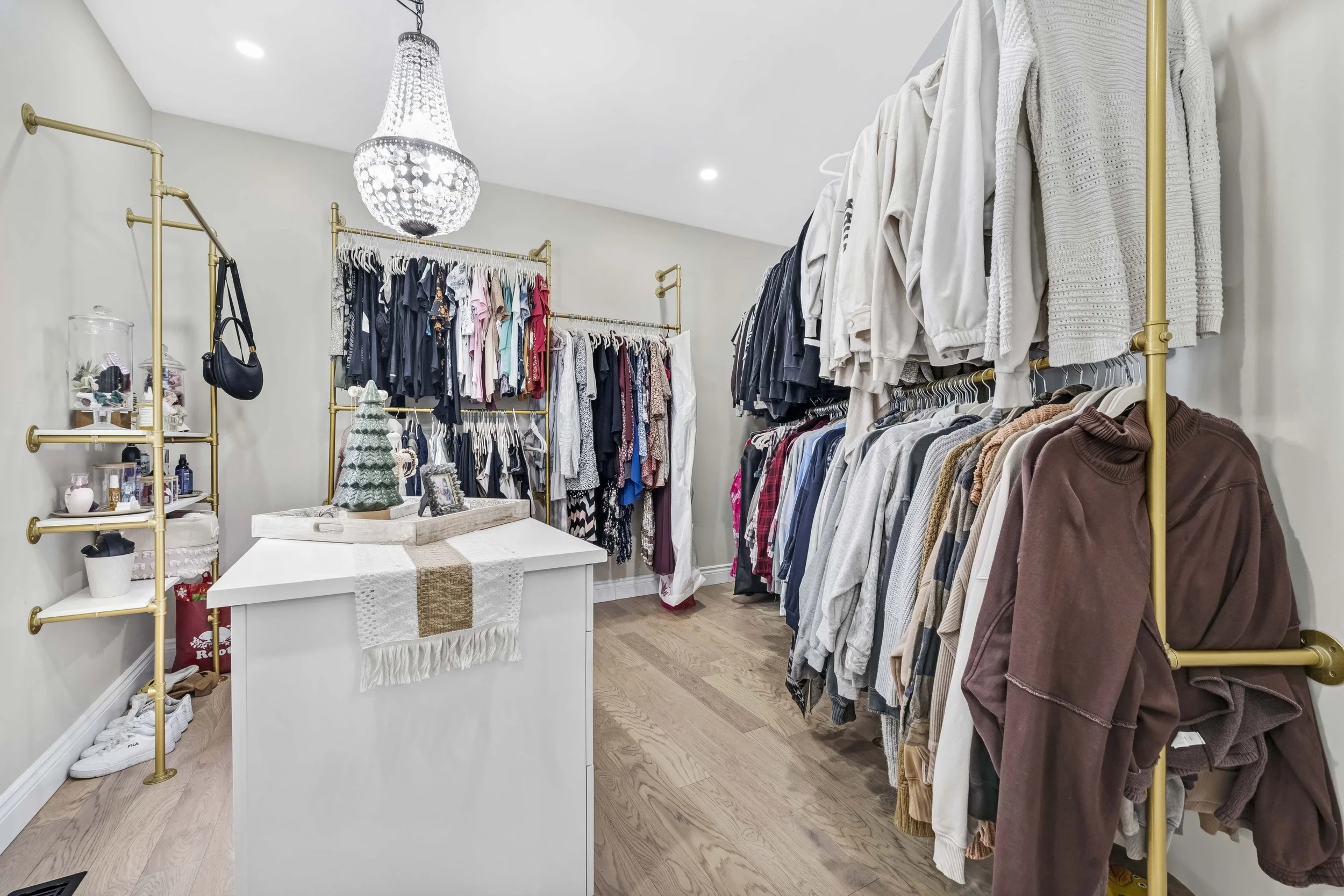photographie immobiliere outaouis  A well-organized walk-in closet with clothing racks filled with various shirts, pants, and sweaters, a white island with decorative items, and gold shelving units on both sides.