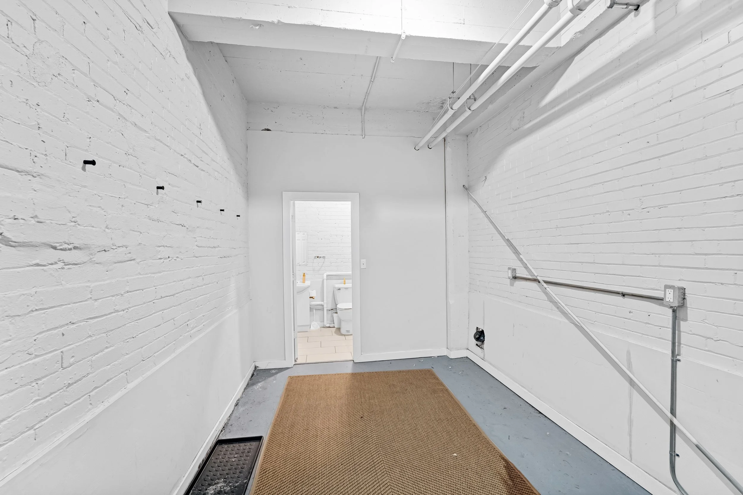 photographie immobiliere outaouis Empty room with white brick walls, a beige rug on a concrete floor, and a doorway leading to a small bathroom with a white toilet and sink.