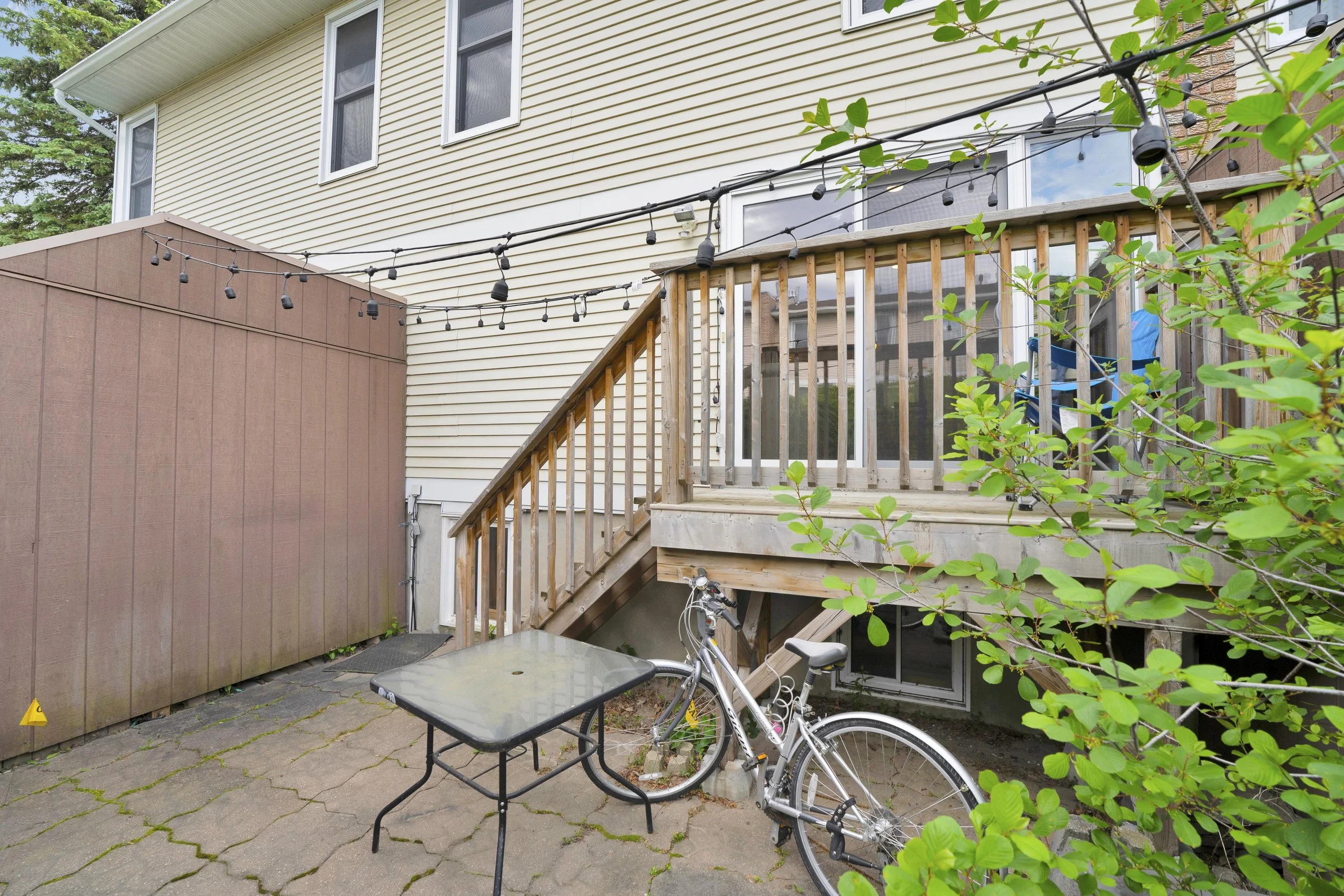 NVISION3D Real Estate Photography Ottawa  Backyard patio area with a bicycle, a small glass table, string lights, and a multi-level deck with wooden stairs. The house has cream siding and white-framed windows.