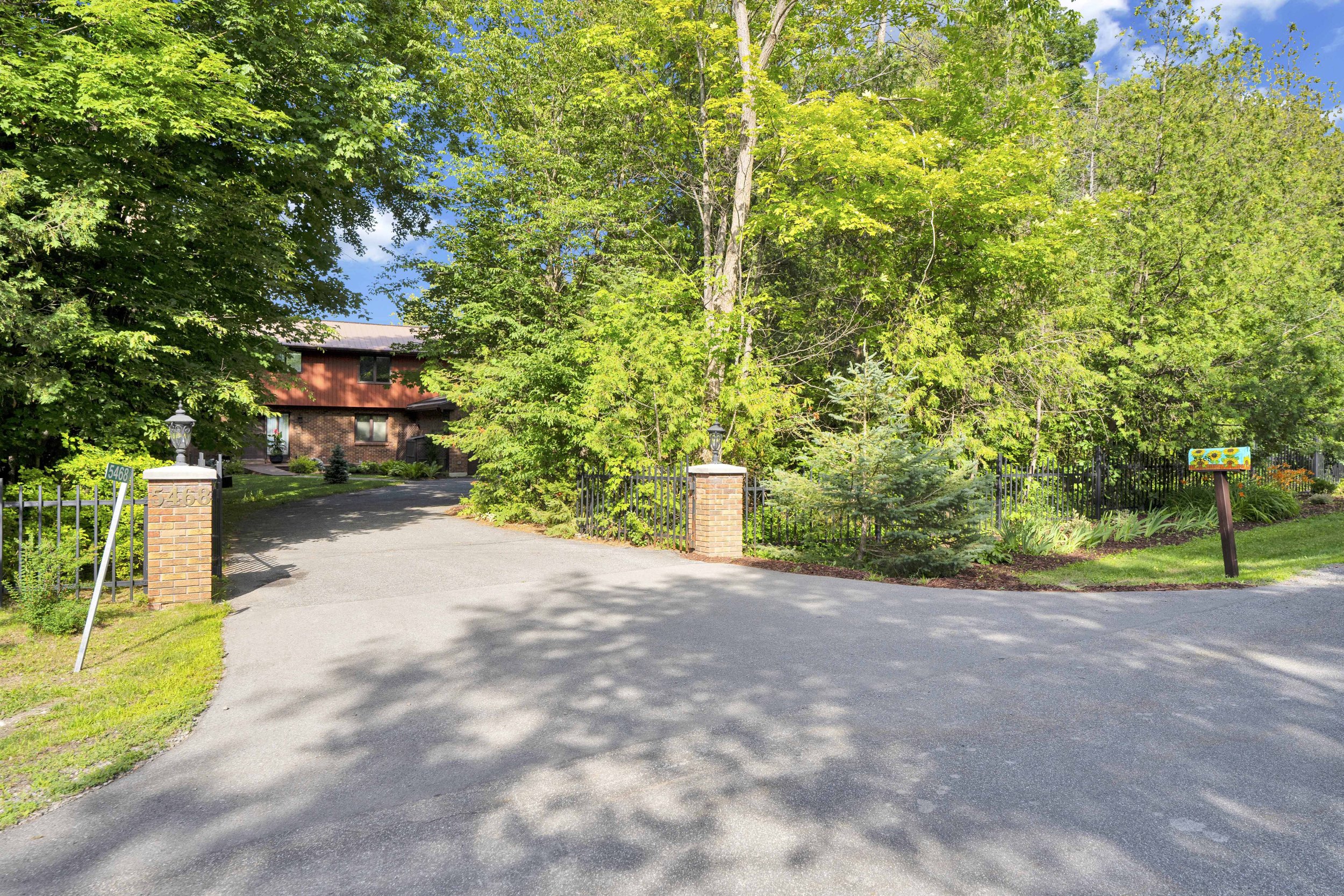 NVISION 3D Real Estate Media  A tree-lined driveway leading to a house, with a brick fence gate, a mailbox, and lush green trees and plants, sunny sky, and shadows on the asphalt.