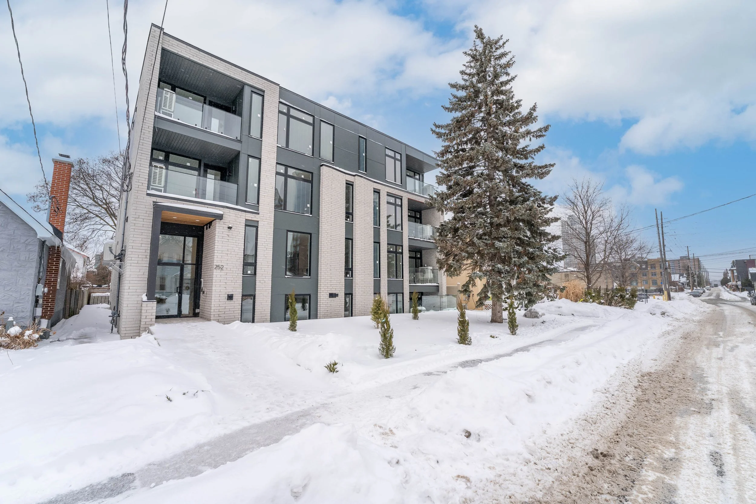 Modern multi-story apartment building with large windows, balconies, and landscaped snow-covered yard, in a snowy winter setting with partly cloudy sky.
