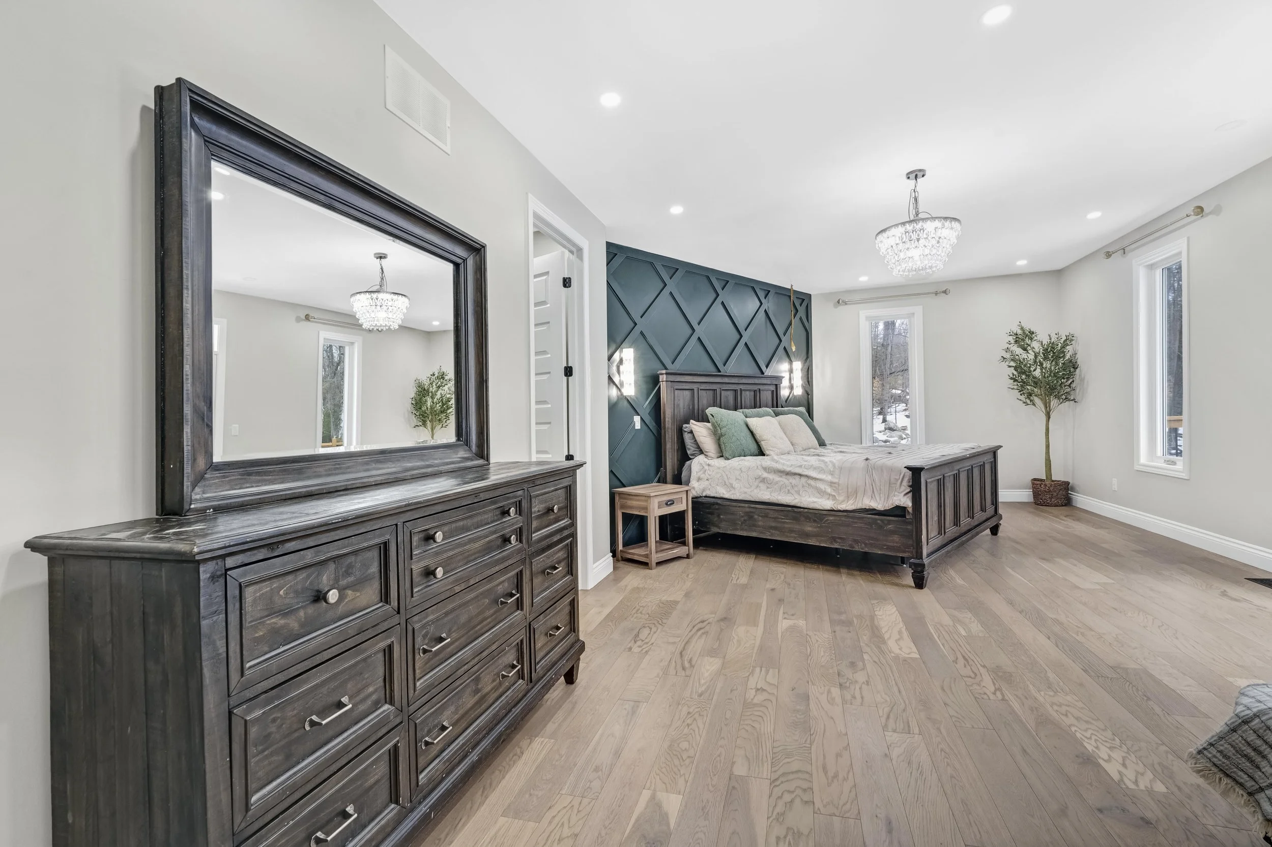 A spacious bedroom with a wooden bed, navy accent wall, and large windows. Includes a wooden dresser with a mirror and a small wooden bedside table. Features a chandelier and a potted plant, with light wood flooring and light-colored walls.