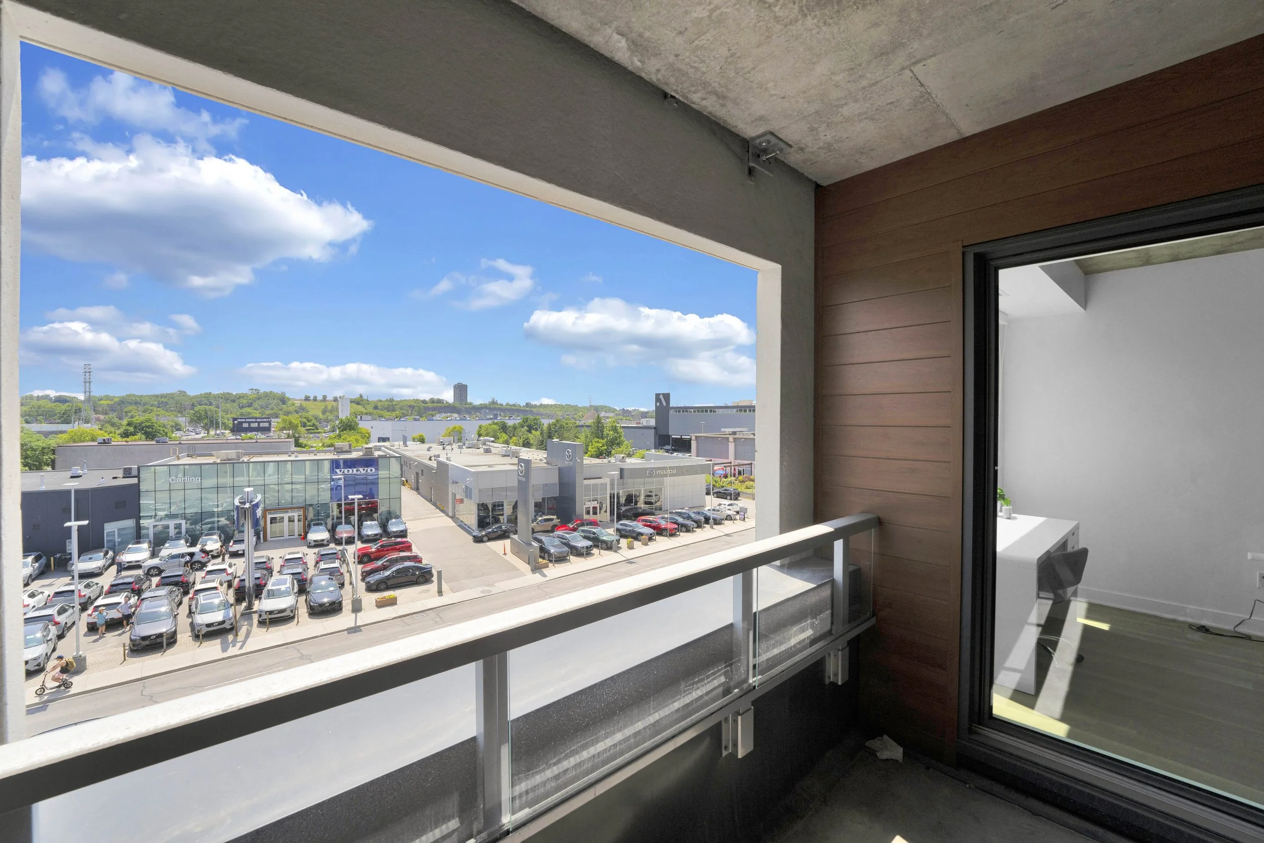 Ottawa Real Estate Videographer NVISION 3D  View from an apartment balcony overlooking a parking lot, commercial buildings, and a green hillside under a partly cloudy sky.