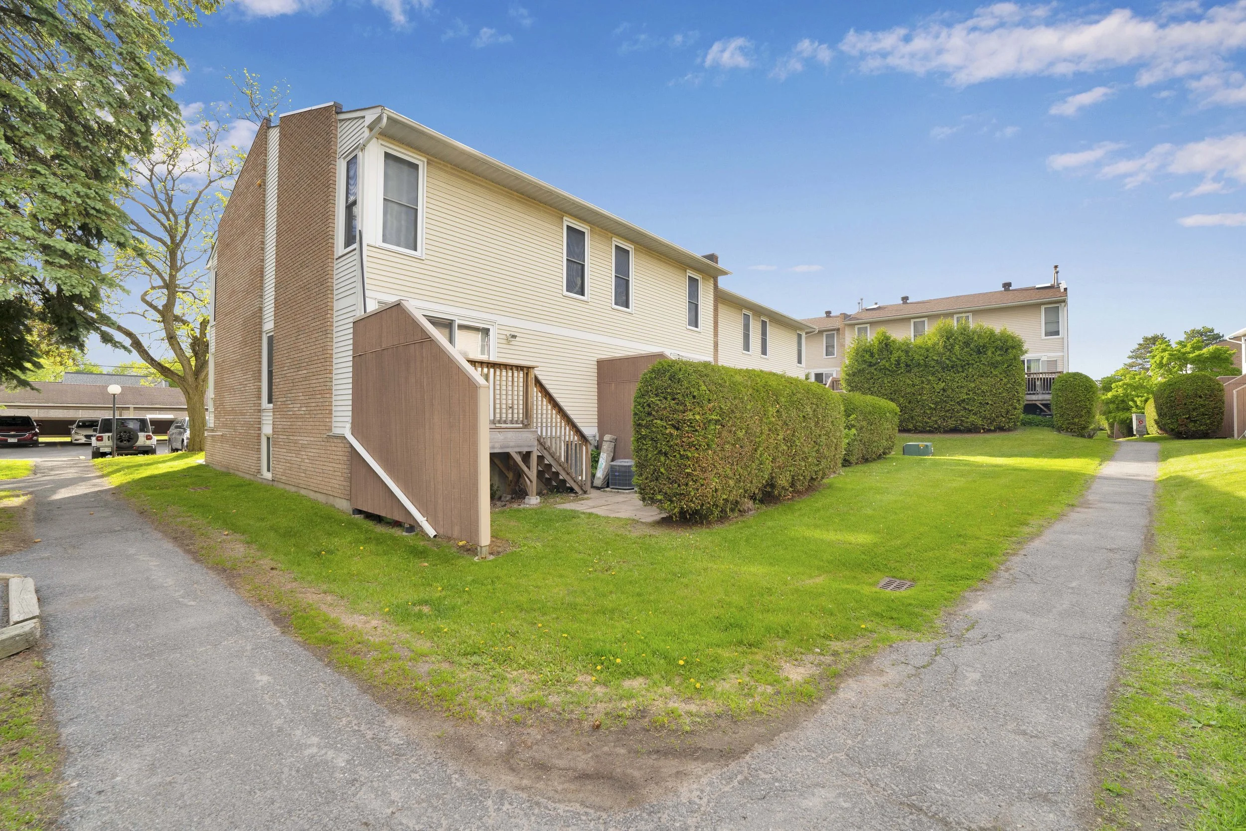 NVISION3D Real Estate Photography Ottawa  A residential apartment complex with beige buildings, a well-maintained lawn, trimmed bushes, trees, and a paved walkway under a blue sky with scattered clouds.