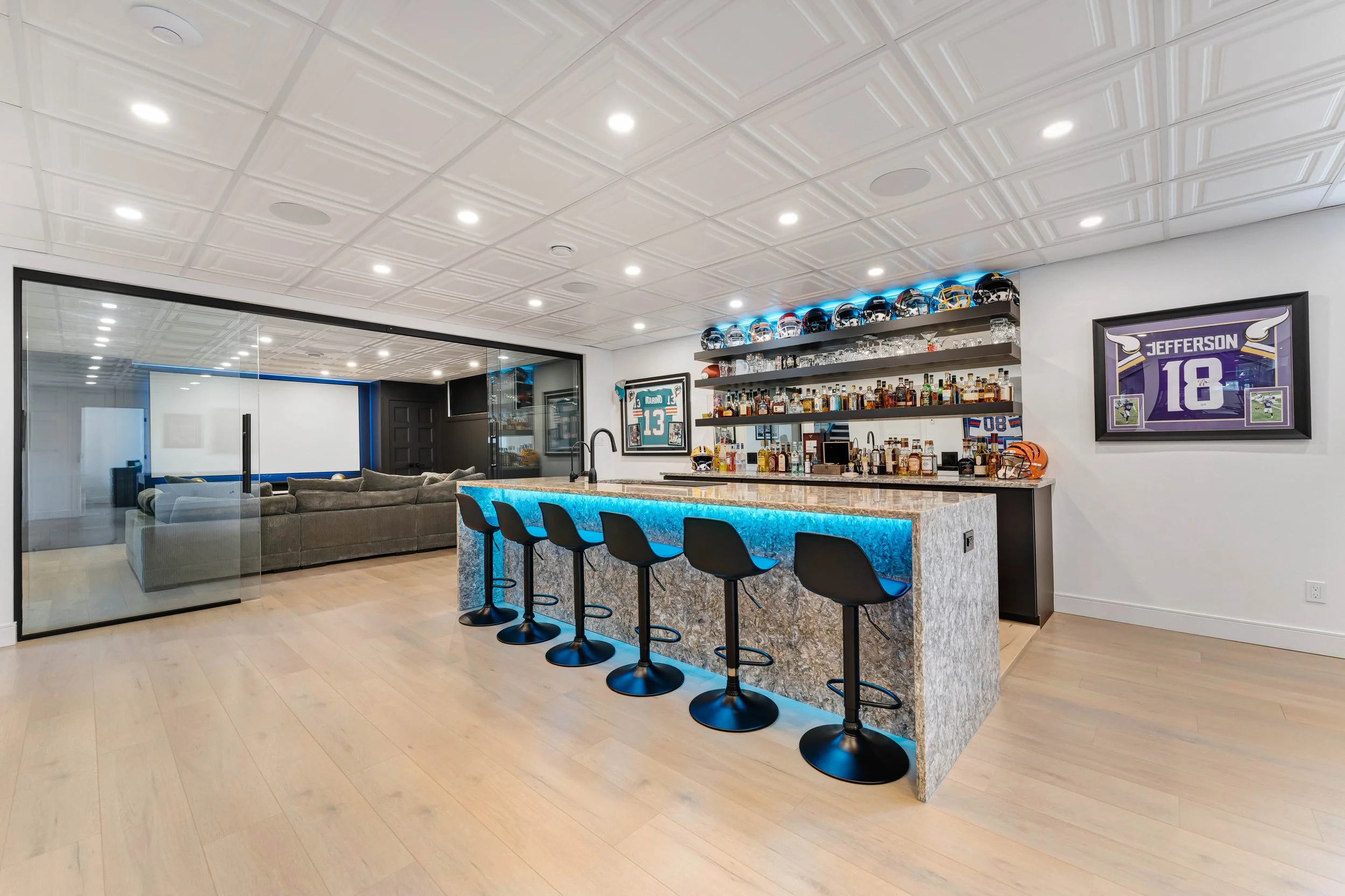 Modern home bar with illuminated counter, black stools, sports memorabilia, and a glass wall separating a lounge area.