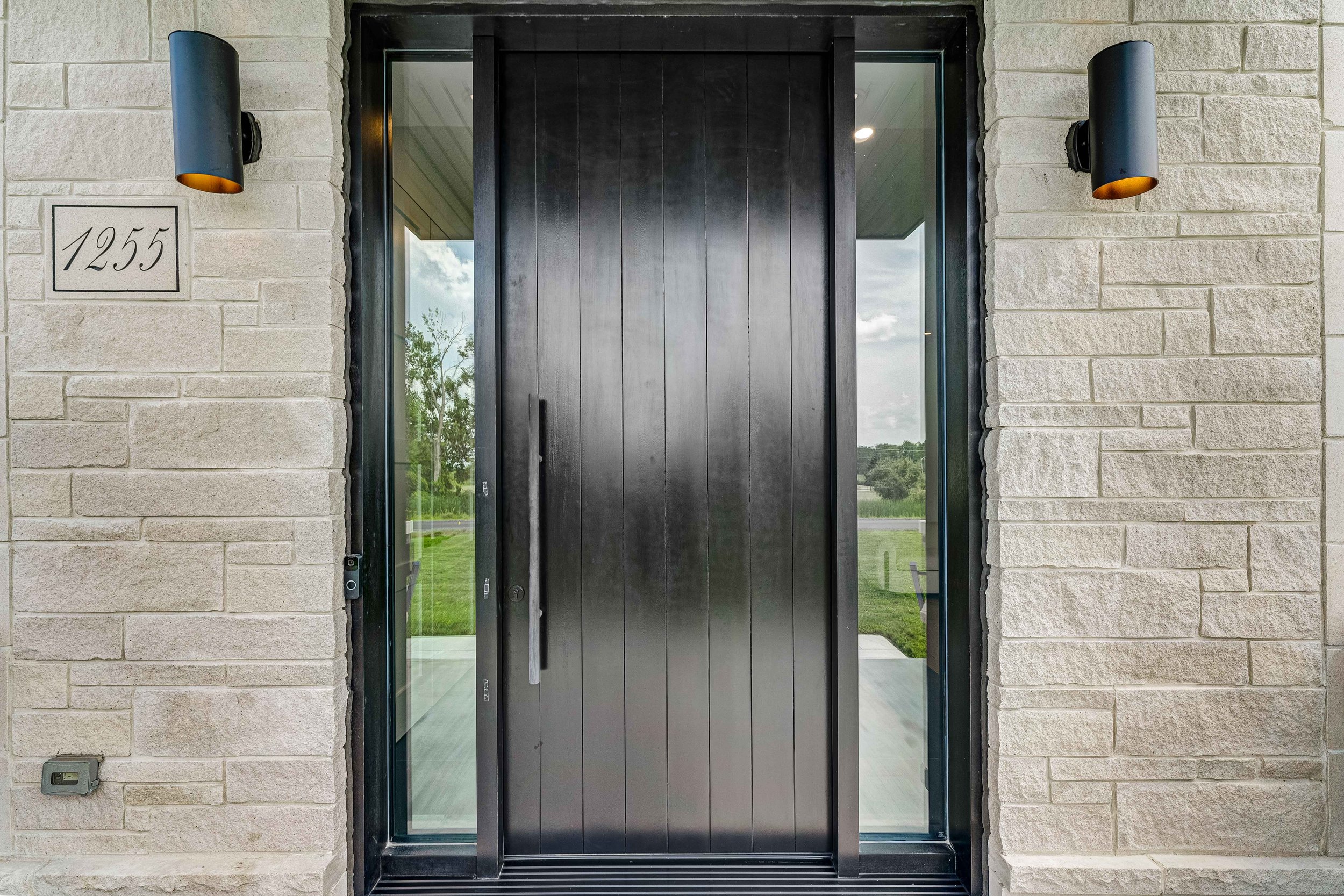 Modern house entrance with a large black door, flanked by sidelights, stone walls, house number 1255, and two black wall sconces.