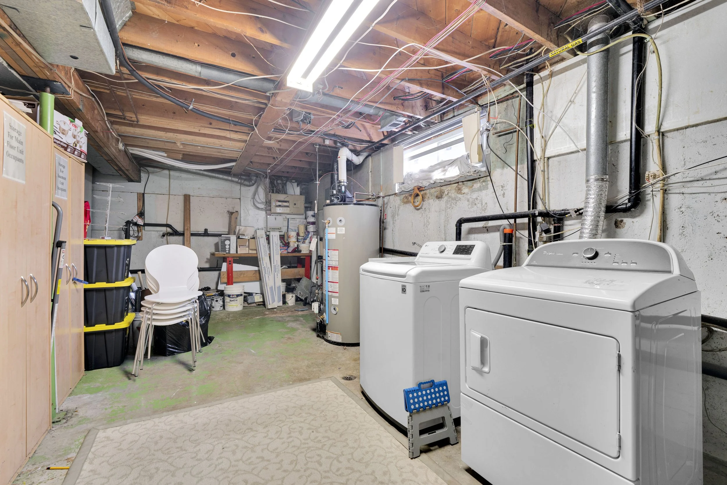 A basement laundry room with unfinished wooden ceiling and exposed pipes. Contains two white appliances, a washing machine, and a dryer. There is a small beige rug, a stack of white chairs, black storage bins, and various tools and supplies stored on