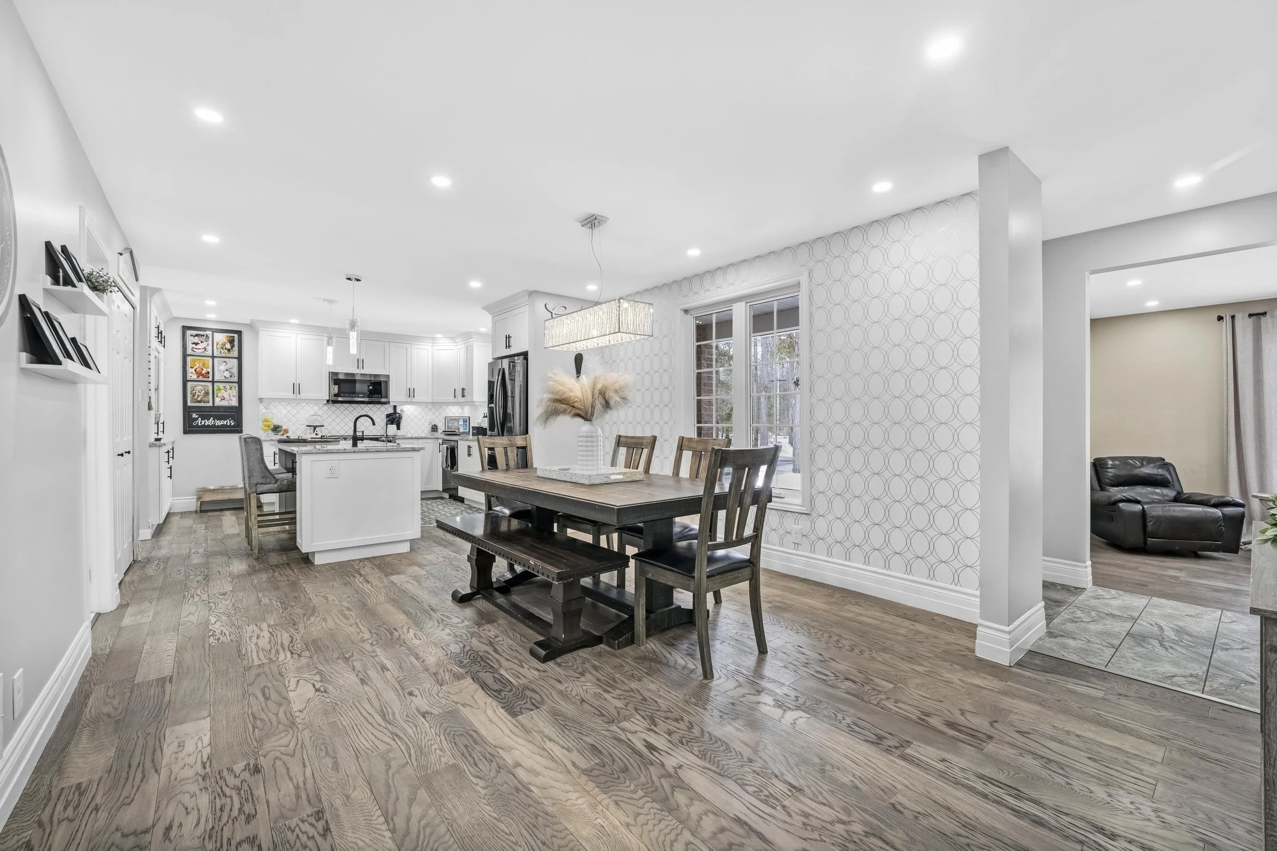 Open-concept kitchen and dining area with wood flooring, white cabinetry, and modern appliances. A dining table with chairs and a decorative vase with pampas grass in the center. Adjacent living space with a black recliner visible through an open doo