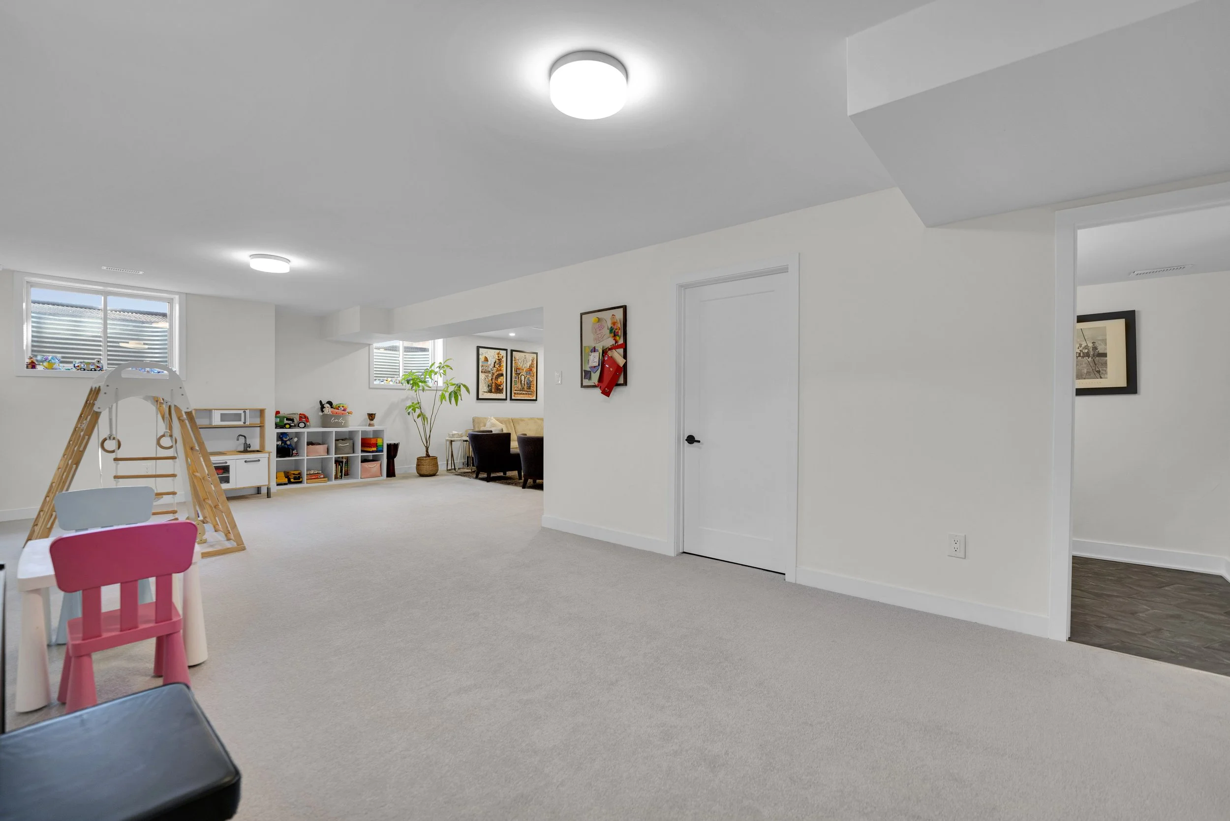 Interior view of a bright, modern room with white walls and ceiling, featuring a children's play area with a pink and white chair, a small table, and shelves with toys, as well as a living space with a beige sofa, artwork on the walls, and large wind