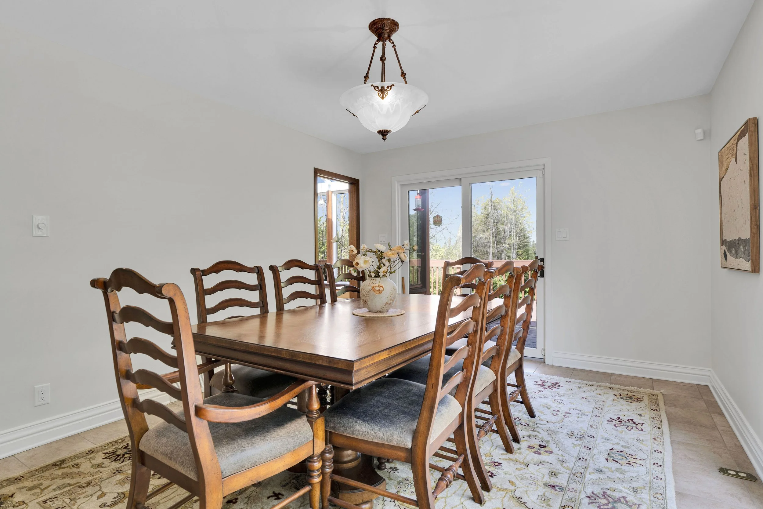 NVISION3D.ca Real Estate Media Ottawa  A dining room with a large wooden table and multiple wooden chairs, a vase with flowers, a ceiling light fixture, and windows revealing an outdoor deck and trees.